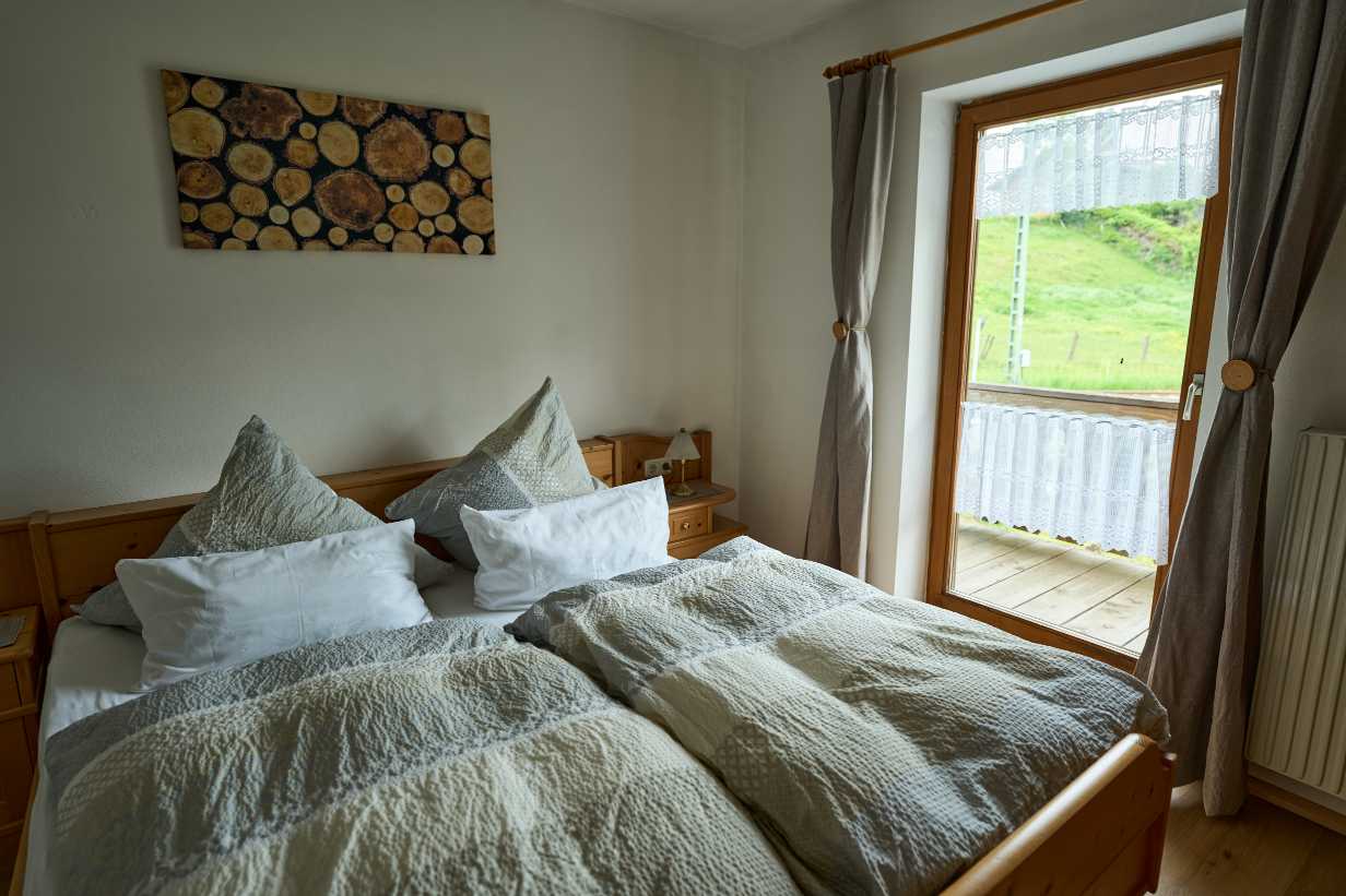 Gemachtes Doppelbett mit grauen und weißen Bettdecken neben einem Fenster mit Blick auf eine grüne Wiese.