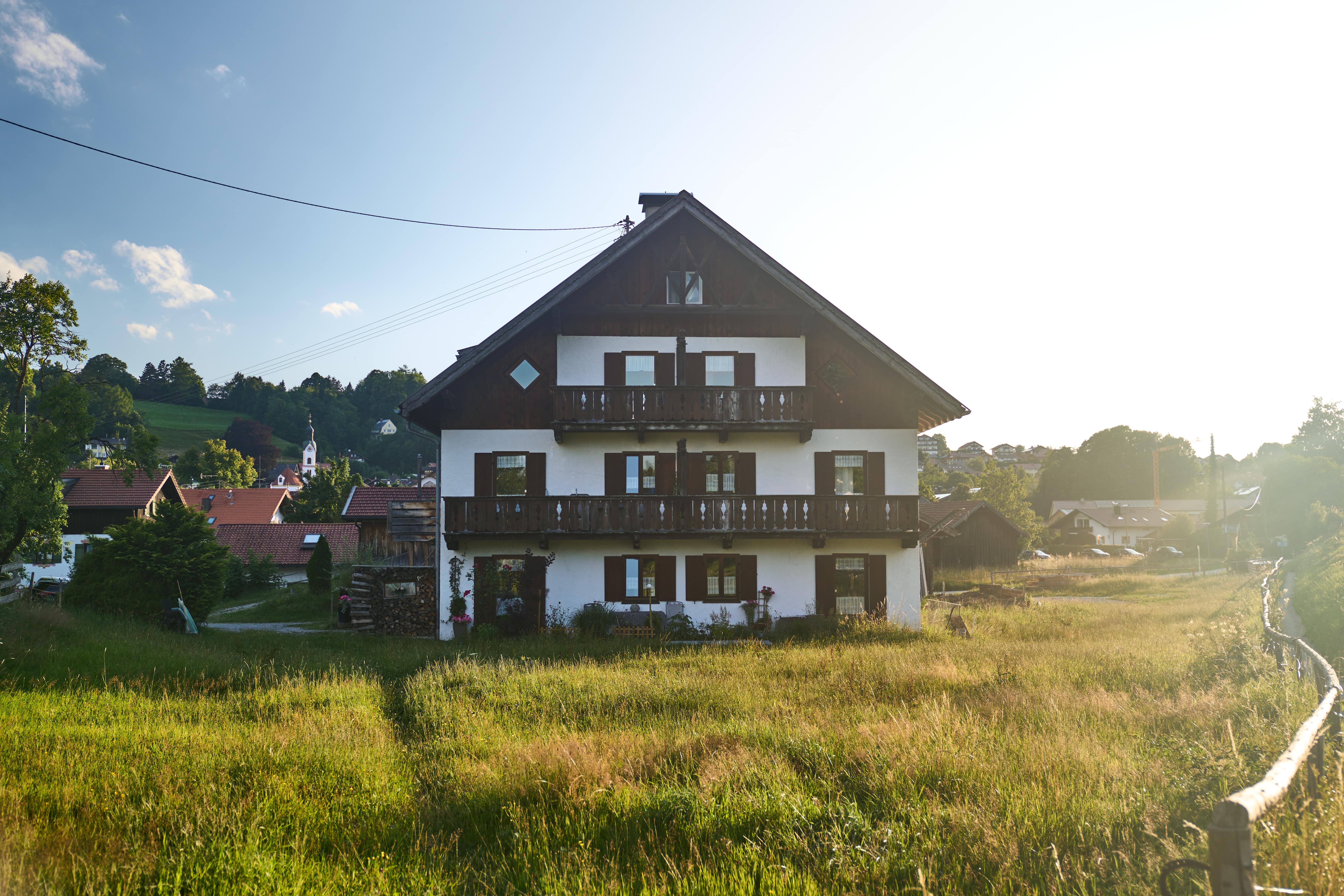 Traditionelles weißes Bauernhaus als Ferienhaus mit dunklen Holzfenstern und Balkonen, umgeben von einer grünen Wiese und umliegenden Häusern im Sonnenlicht in den Ammergauer Alpen.