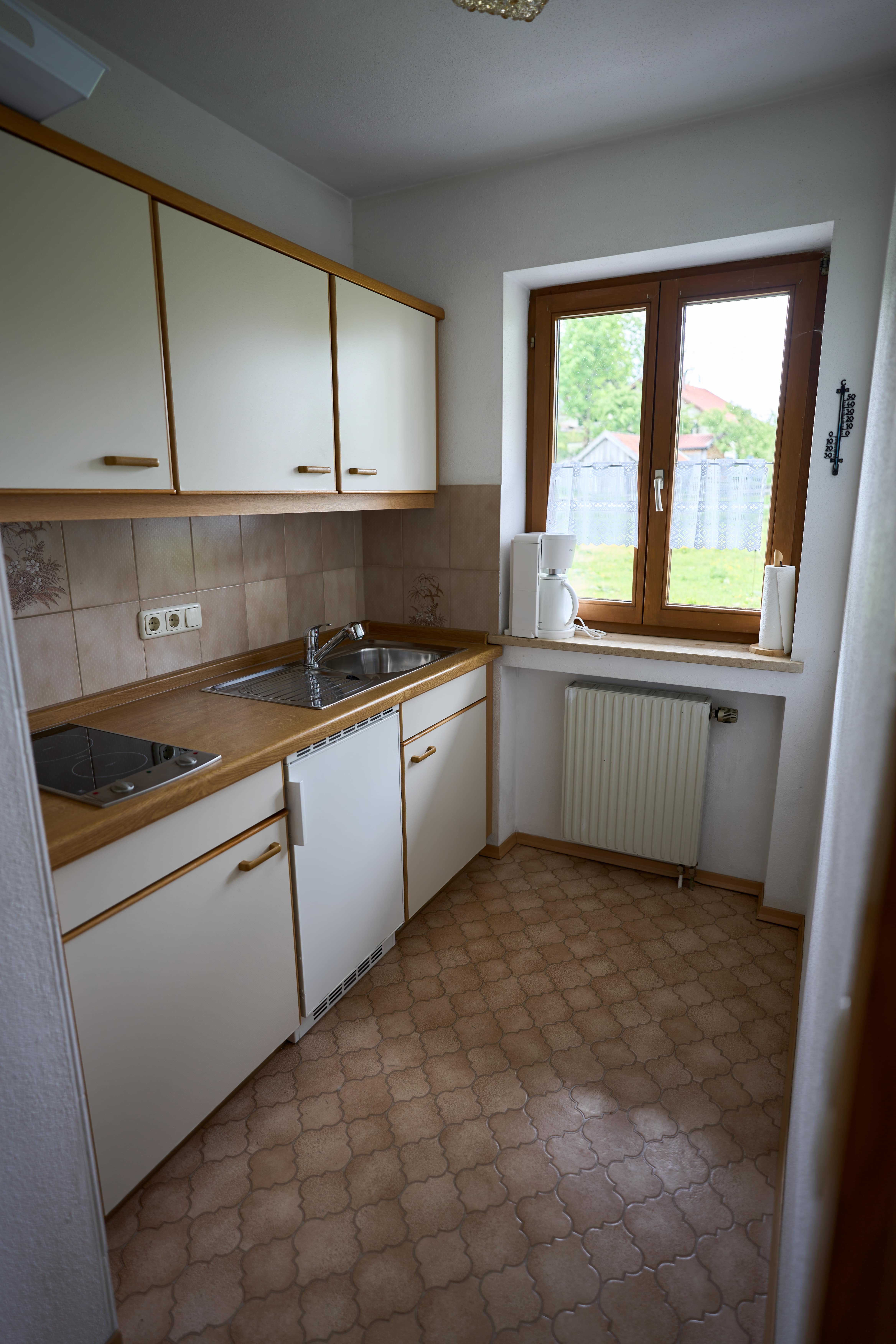 Kleine Küche mit hellen Schränken, Spüle, Kochfeld, Kaffeemaschine auf der Fensterbank und Fenster mit Blick auf grünen Garten.