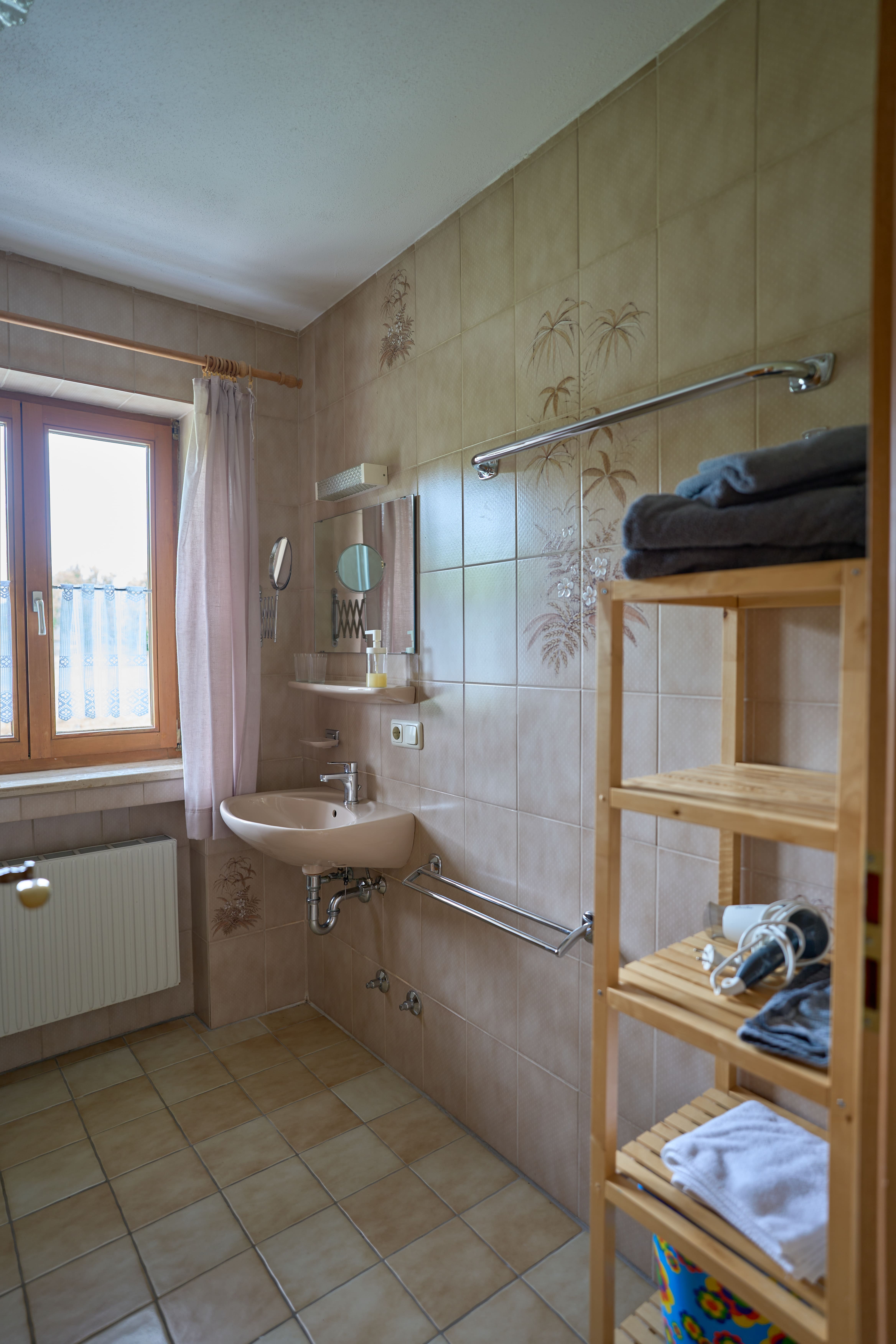 Kleines Badezimmer mit beige gefliesten Wänden, Waschbecken, Spiegel, Fenster mit Vorhängen und einem Holzregal mit Handtüchern und Haartrockner.