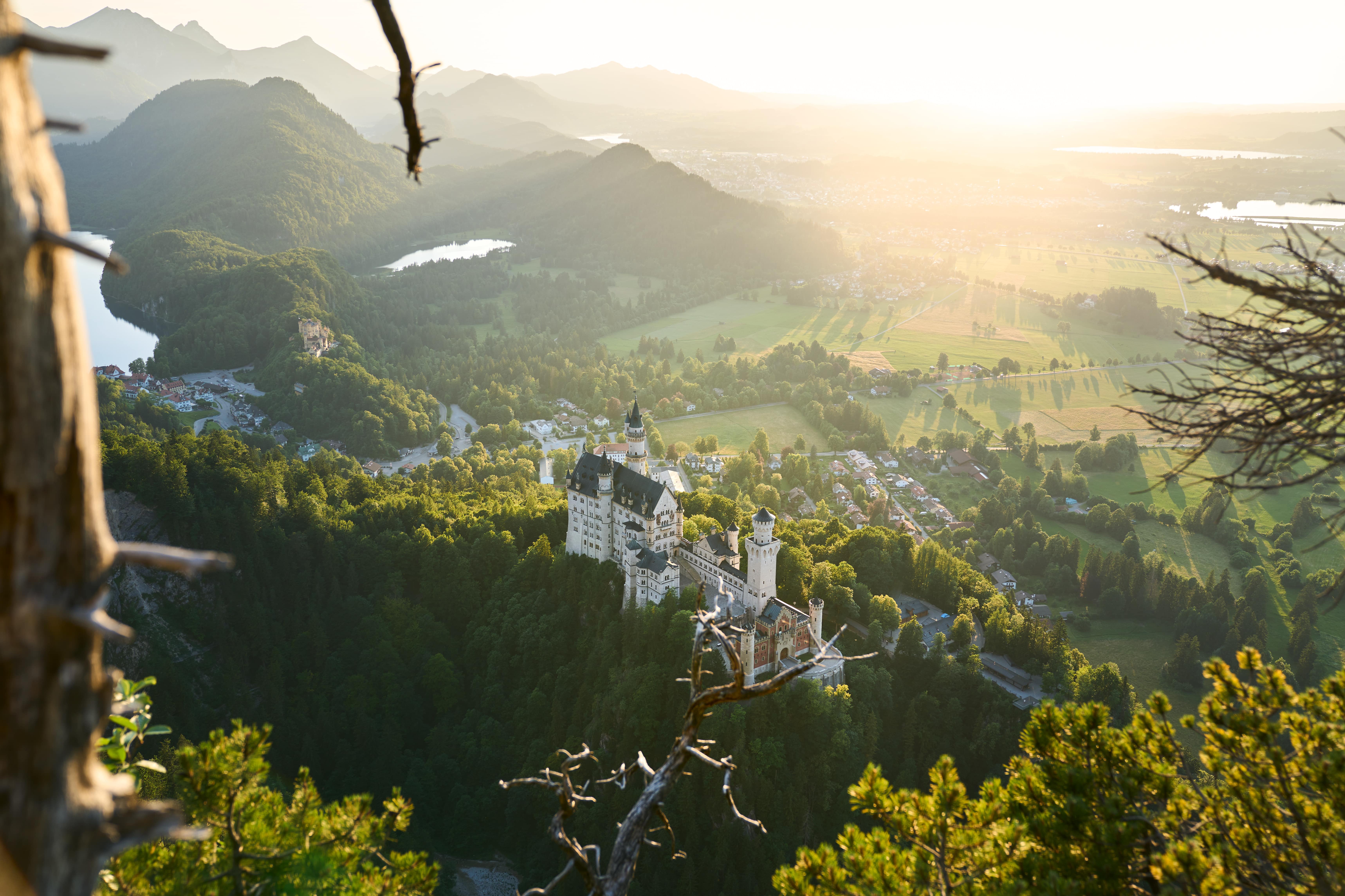 Luftaufnahme von Schloss Neuschwanstein, umgeben von Wald und Bergen bei Sonnenuntergang.