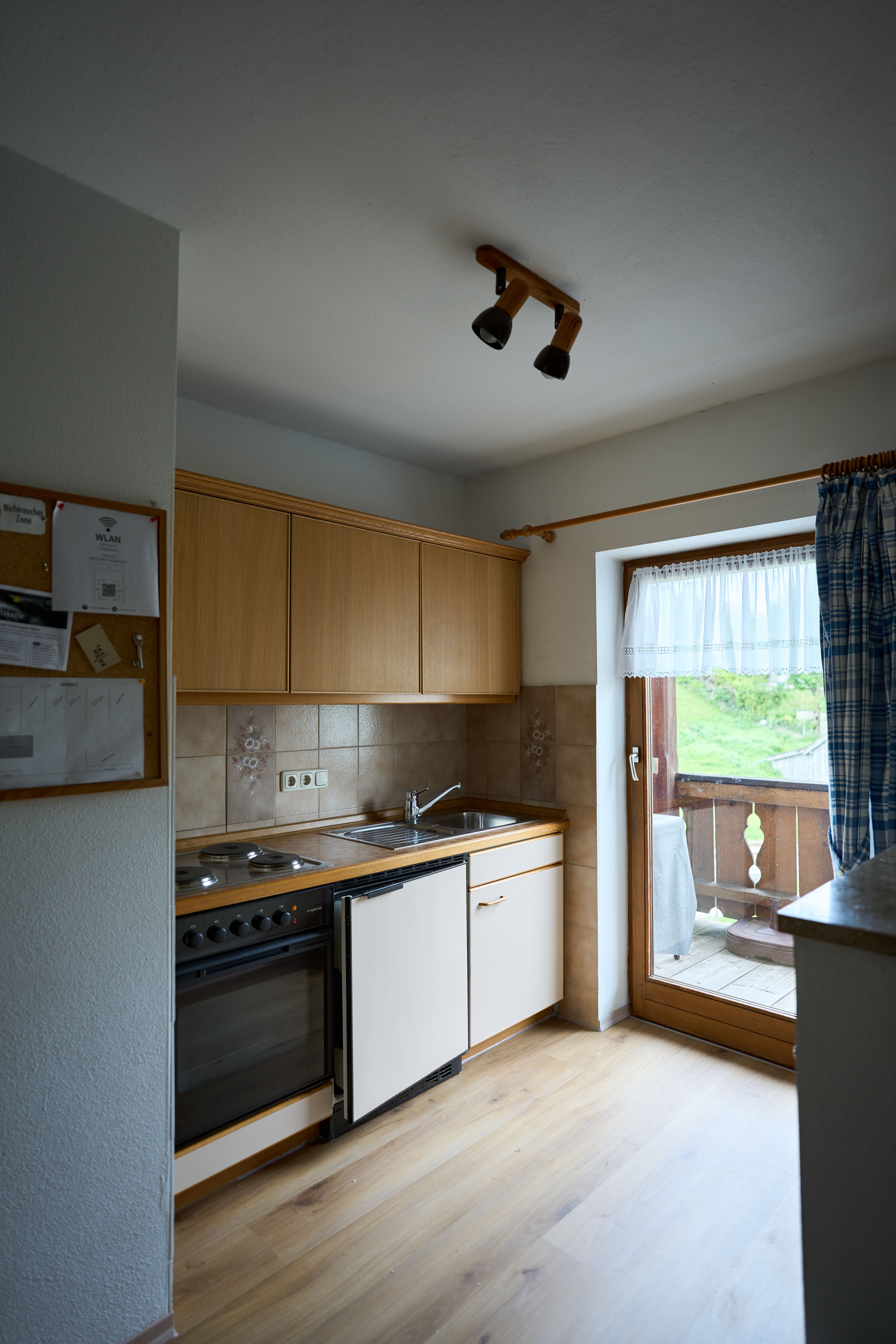 Kleine Küche mit Holzschränken, Herd, Spüle und Glastür, die auf einen Balkon mit grüner Landschaft führt.