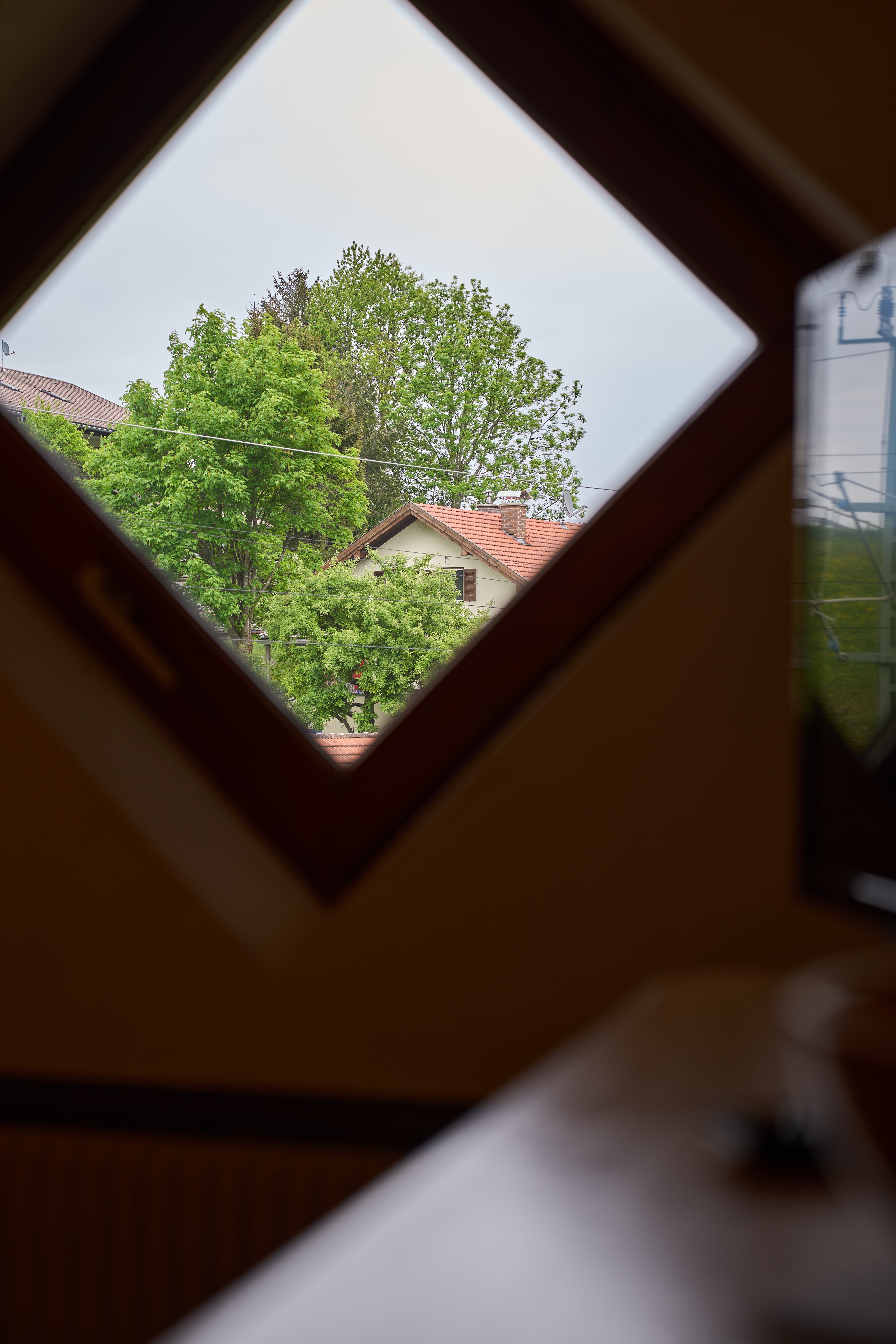 Blick durch ein quadratisches Dachfenster auf ein Haus mit rotem Ziegeldach und grüne Bäume bei bewölktem Himmel.