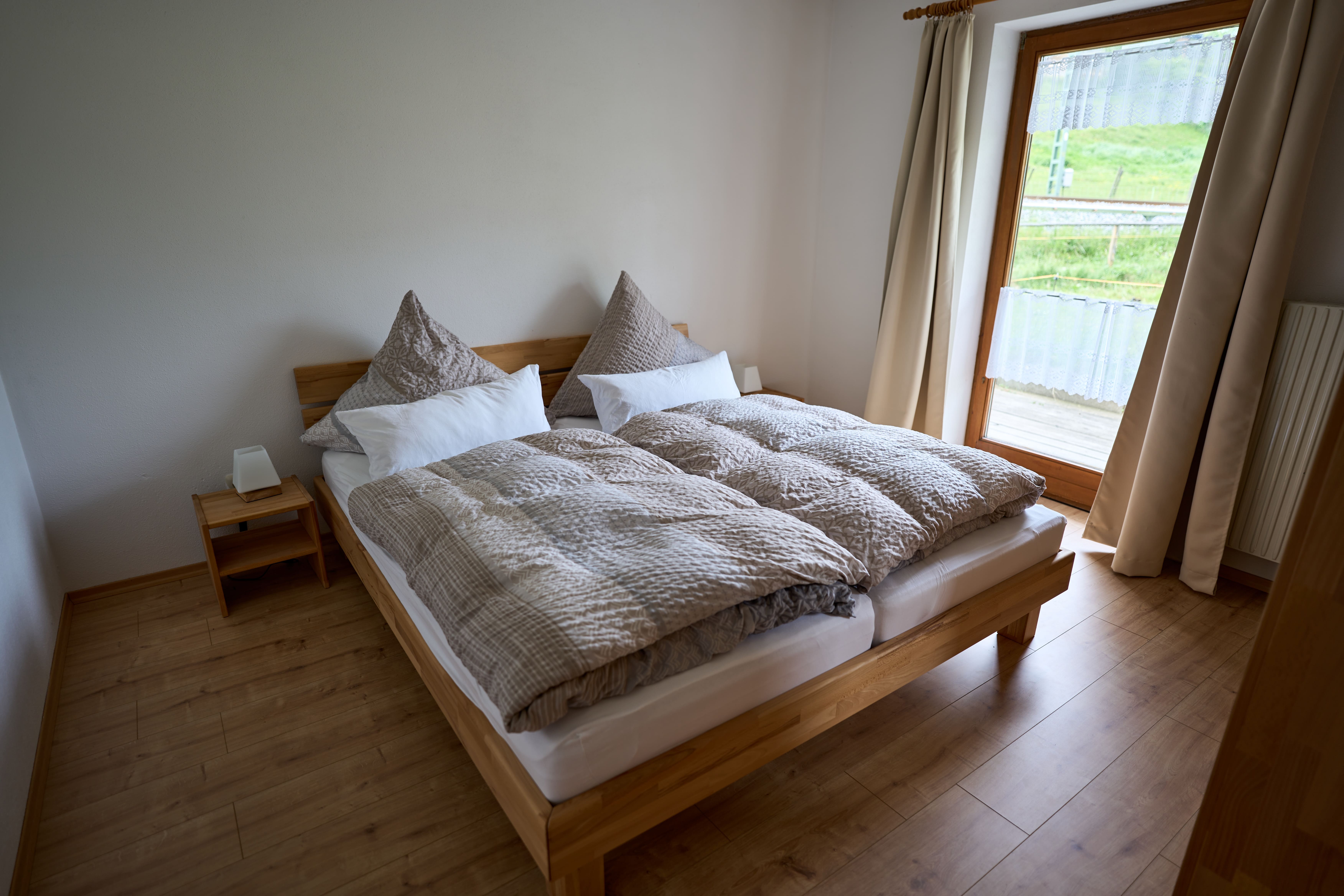Gemütliches Schlafzimmer mit einem Holzbett, braunen Bettdecken und Kissen sowie einem großen Fenster mit Blick auf eine grüne Wiese.