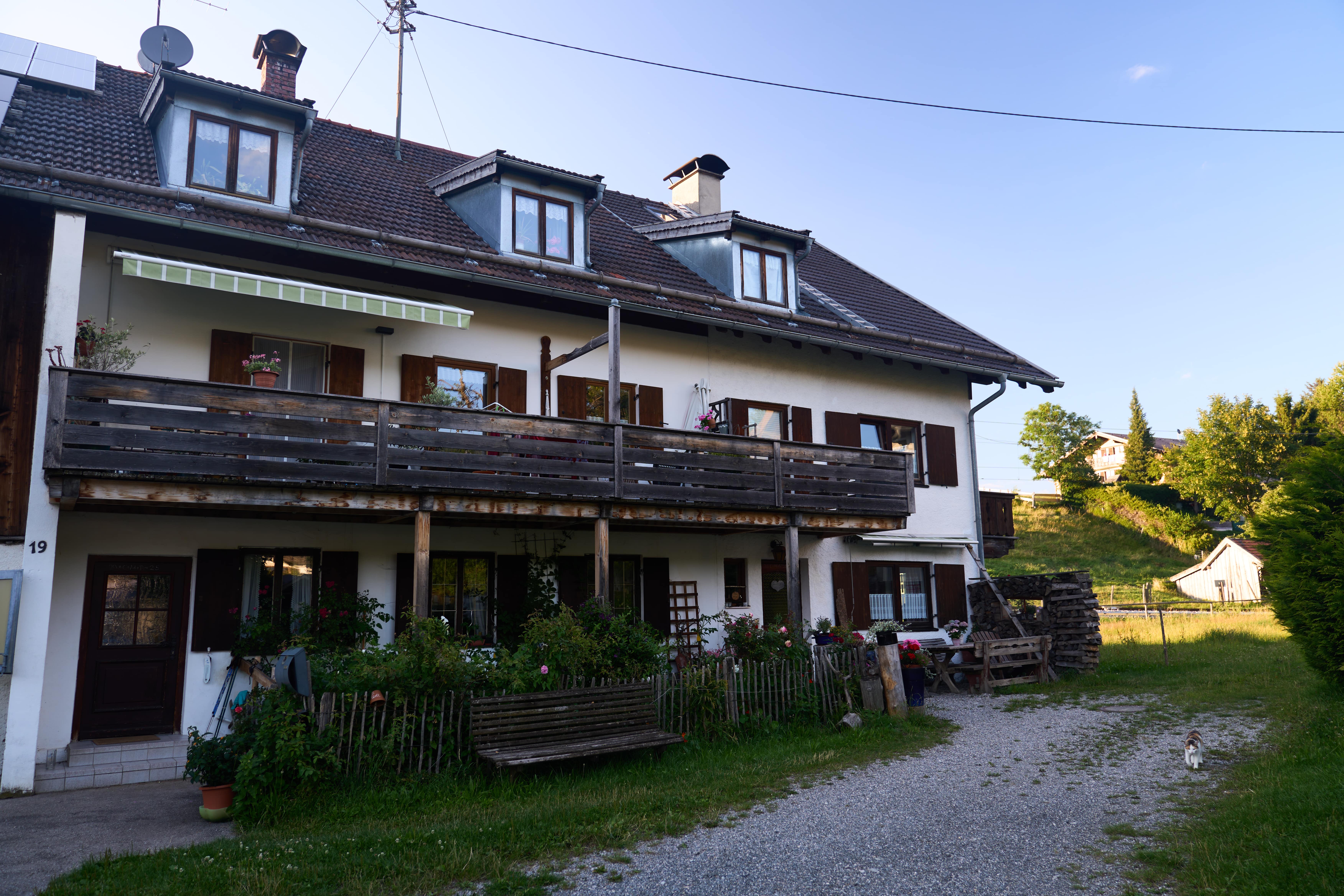 Traditionelles Bauernhaus mit Holzbalkon, Garten mit Blumen, Holzbänken und einer Katze auf einem Kiesweg im Abendlicht.