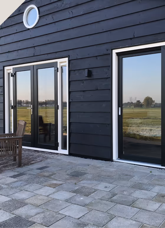Zwart houten huis met grote glazen deuren en een ronde raam, met uitzicht op een groen veld en een betegeld terras.