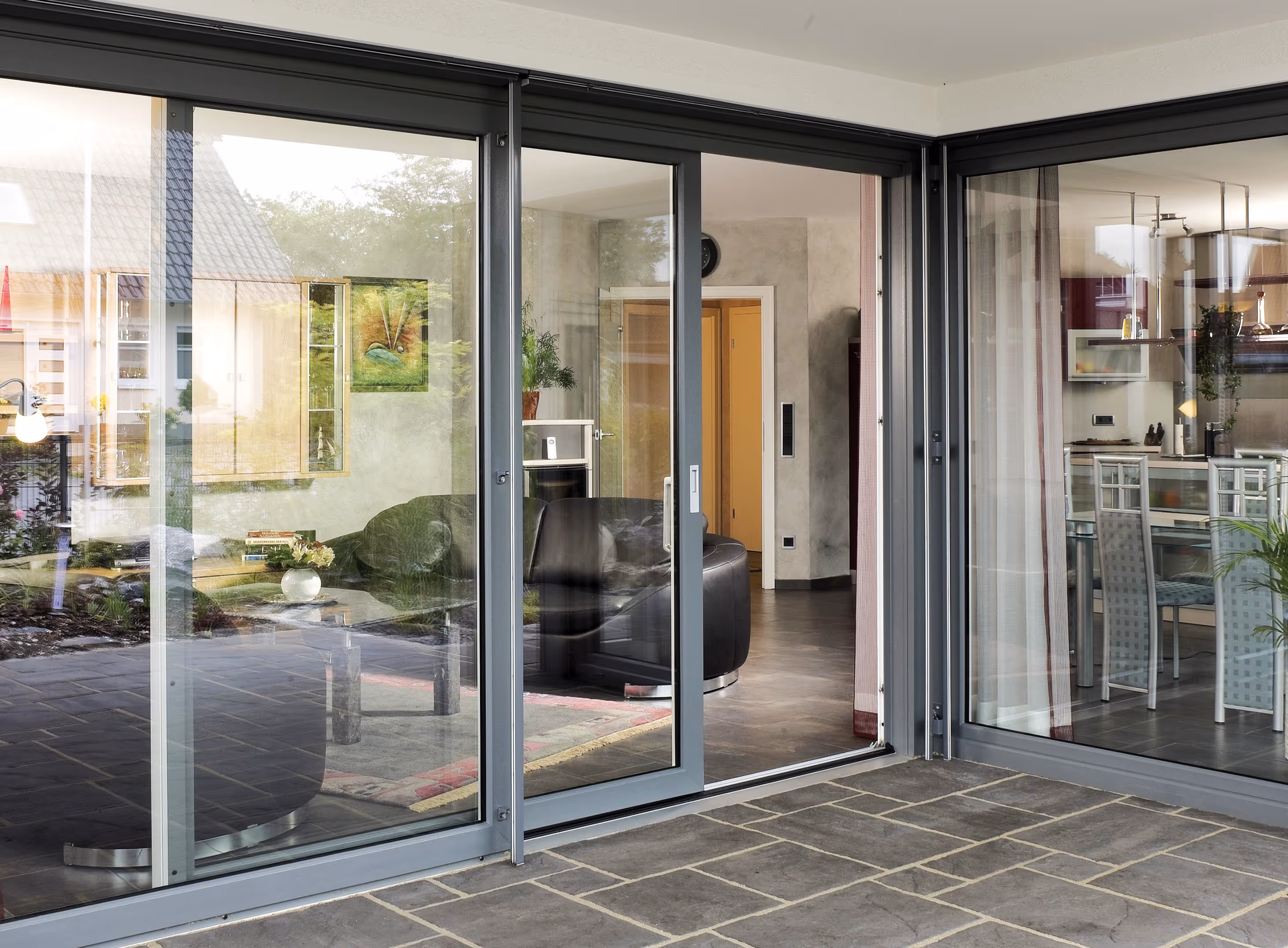 Moderne schuifdeur van glas met uitzicht op een woonkamer met zwart leren bank en een eetkamer met een glazen tafel en witte stoelen.