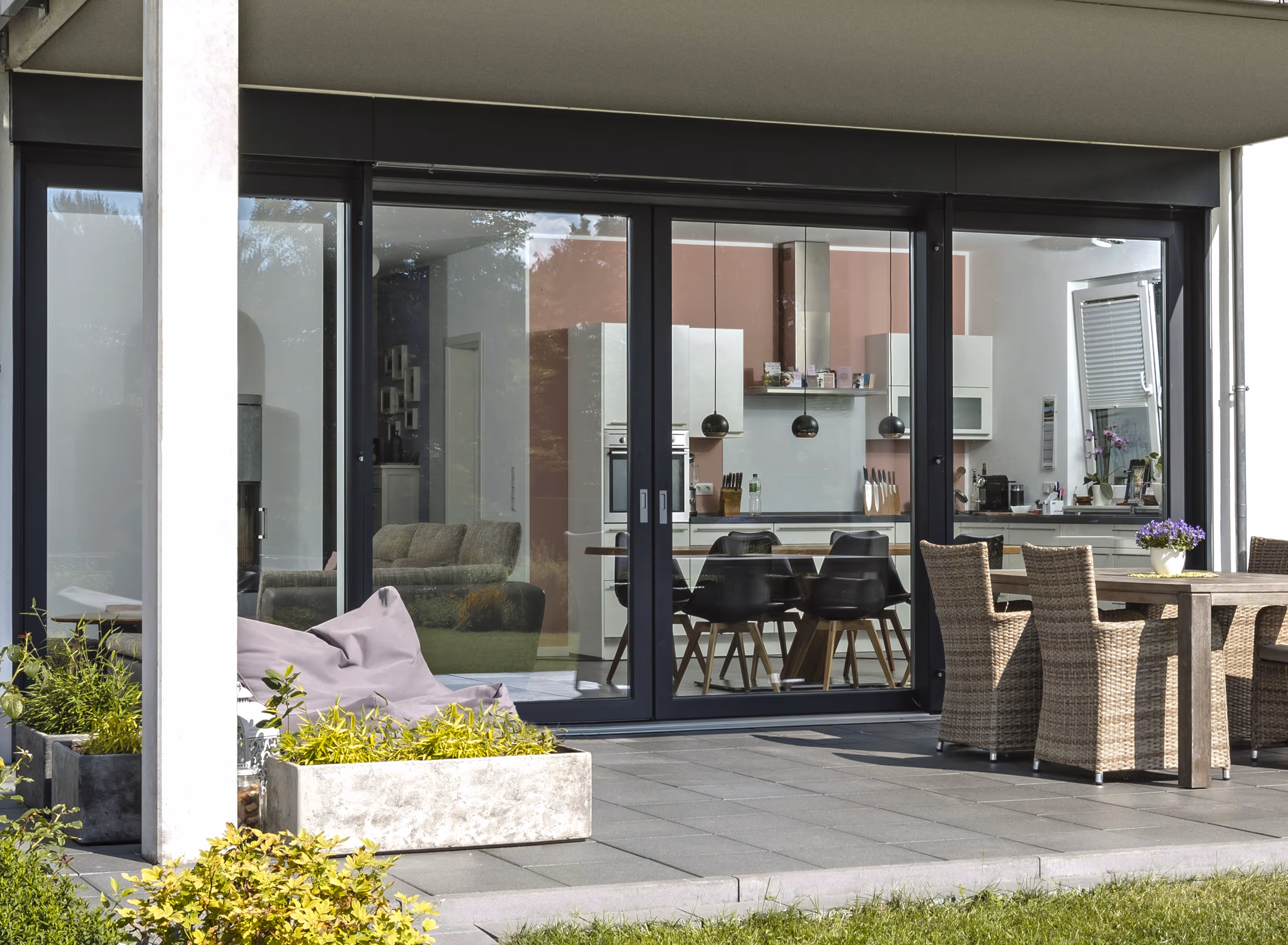 Moderne patio met grijze tegels, druipstoel, plantenbakken en aanliggende woning met grote glazen schuifdeuren naar een keuken en eetkamer.