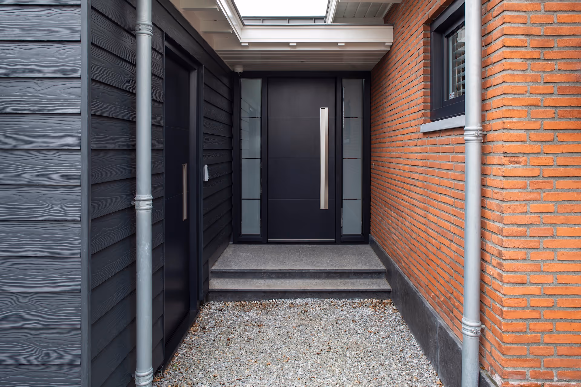 Moderne voordeur van een huis met zwarte houten en bakstenen gevels, grijze stenen traptreden en grindpad.