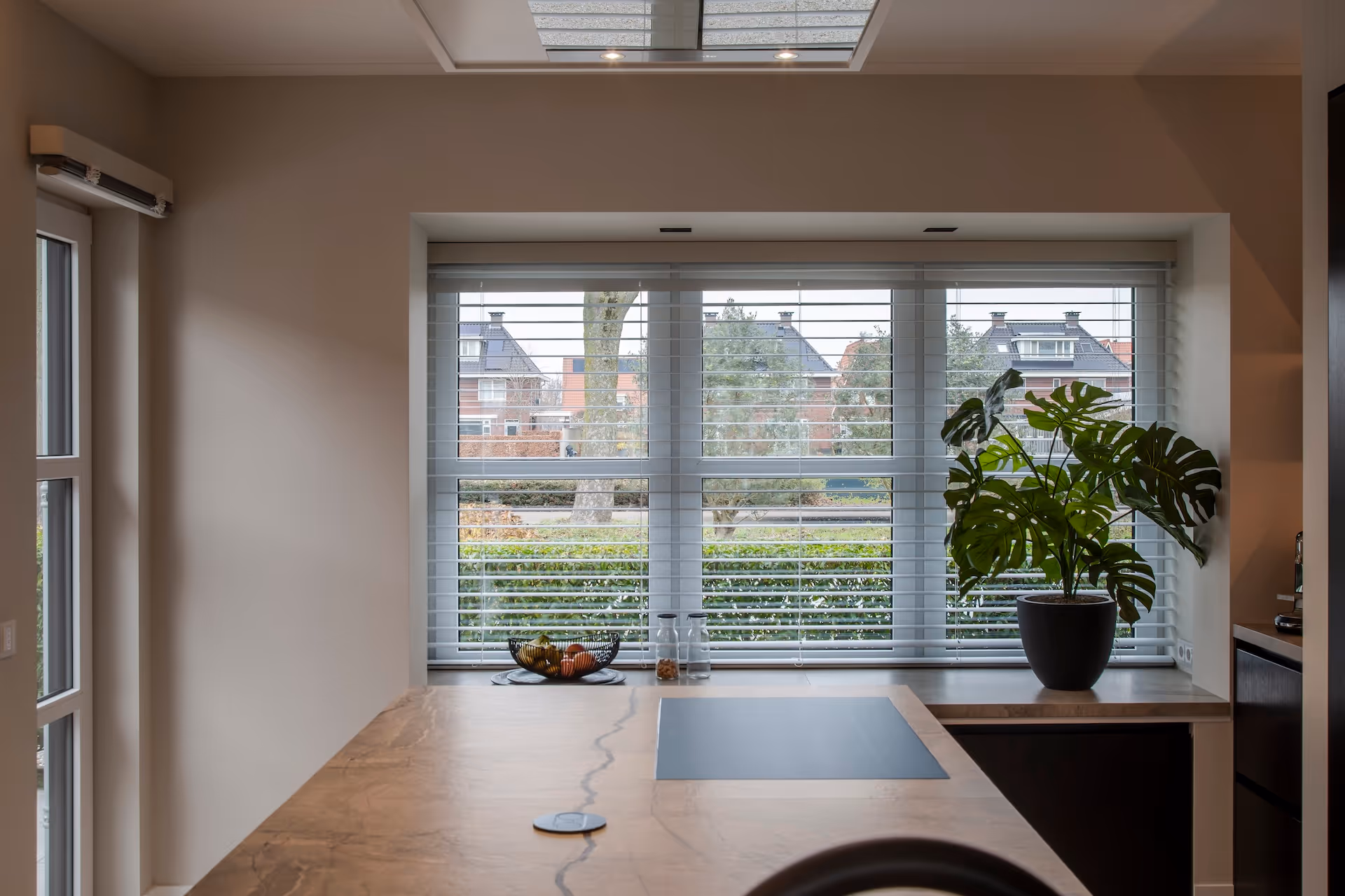 Moderne keuken met houten aanrechtblad, glazen fruitschaal, plantenpot en groot raam met jaloezieën.