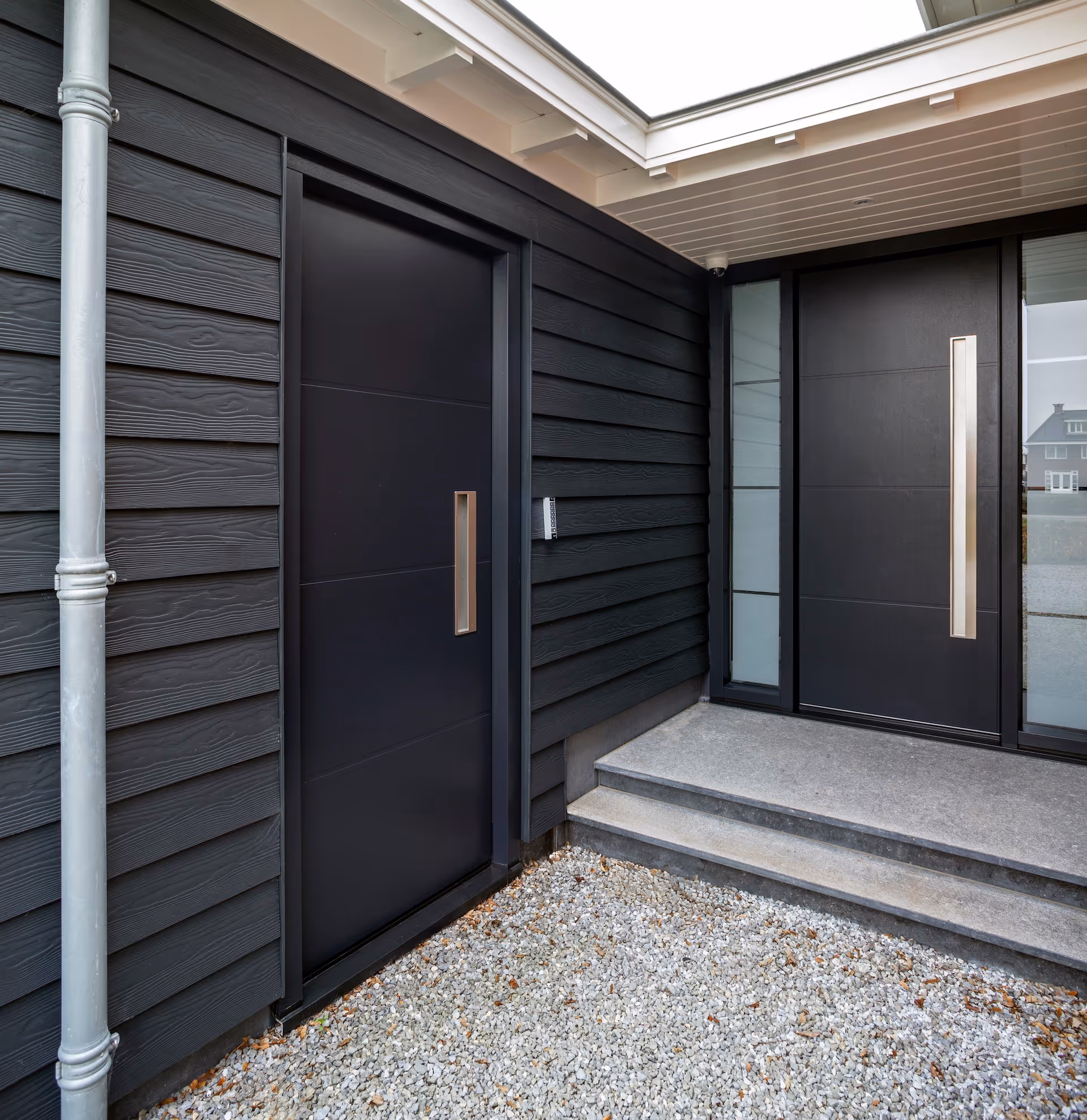 Hoek van een modern huis met zwarte houten gevelbekleding en twee zwarte voordeuren met metalen handgrepen, één met twee kleine treden ernaartoe.