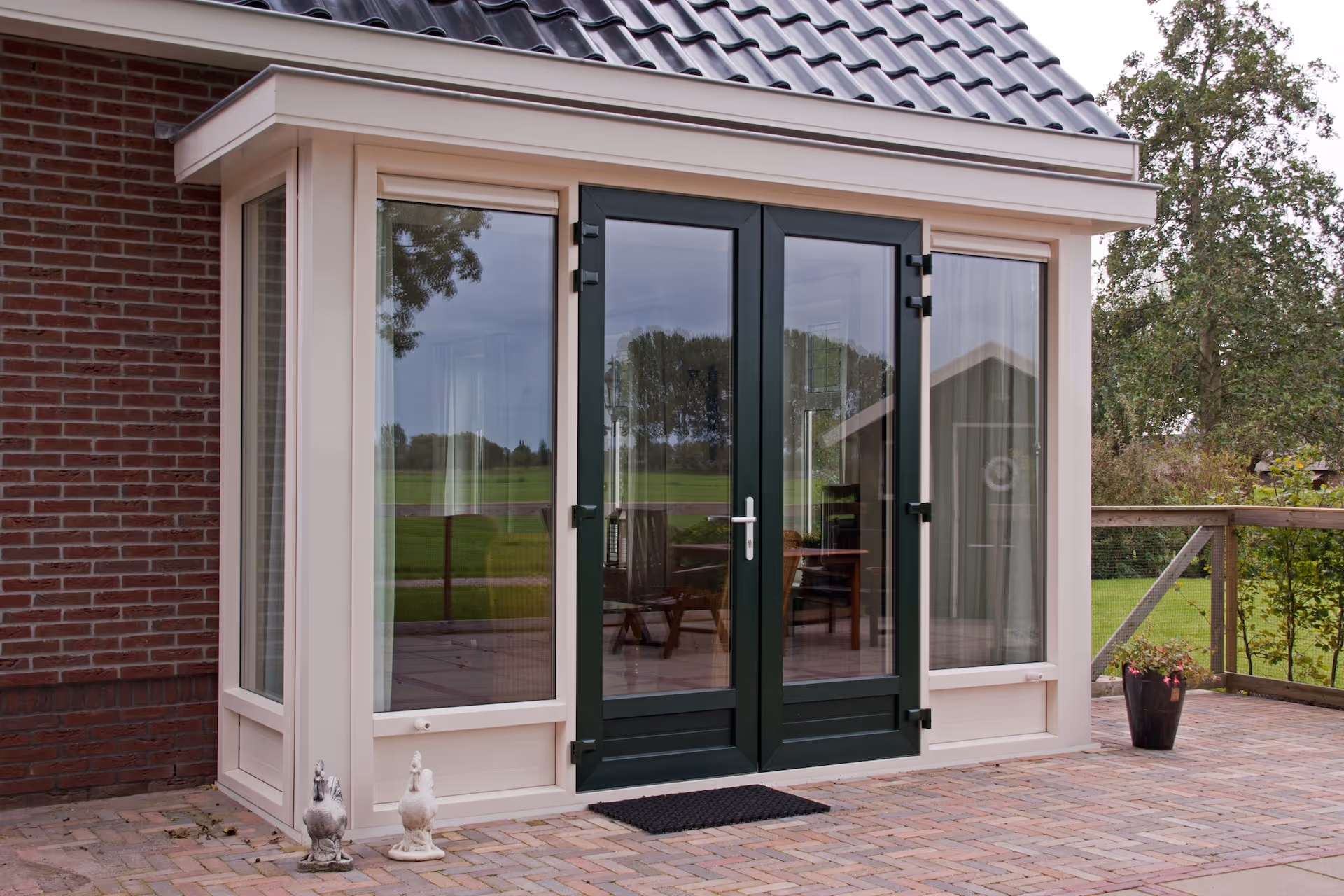 Een huis met donkere dubbele glazen deuren en ramen, met twee witte beeldjes aan de linkerkant en een bloempot rechts op een bakstenen terras.