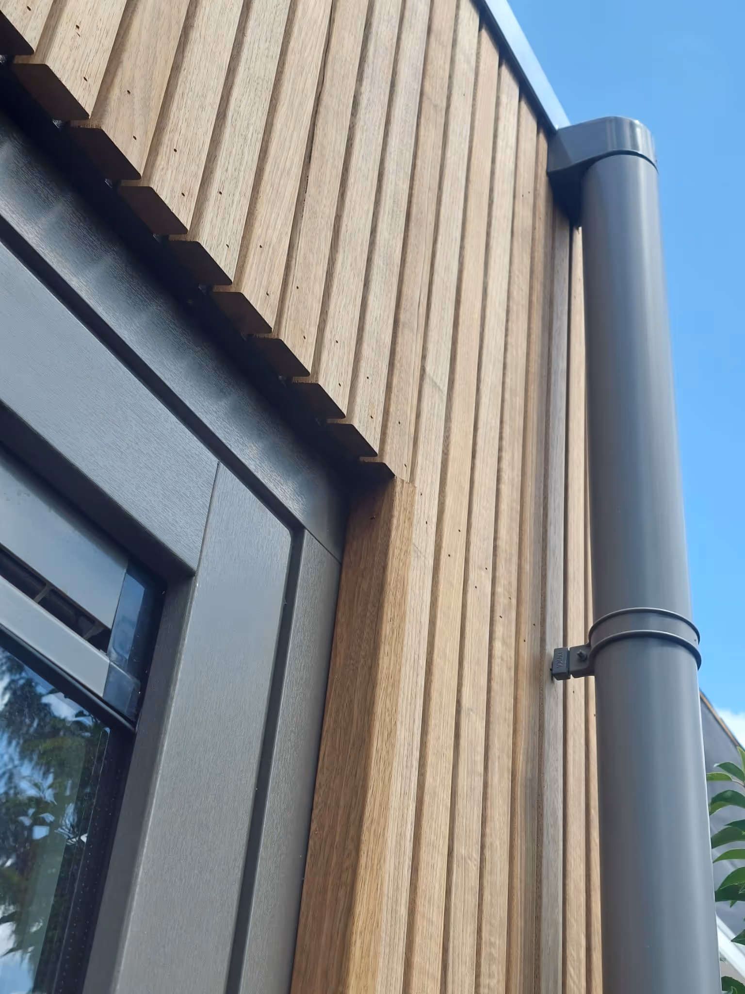 Dichtbij van een huis met verticaal houten gevelbekleding naast een zwarte deur en een grijze regenpijp tegen een blauwe lucht.