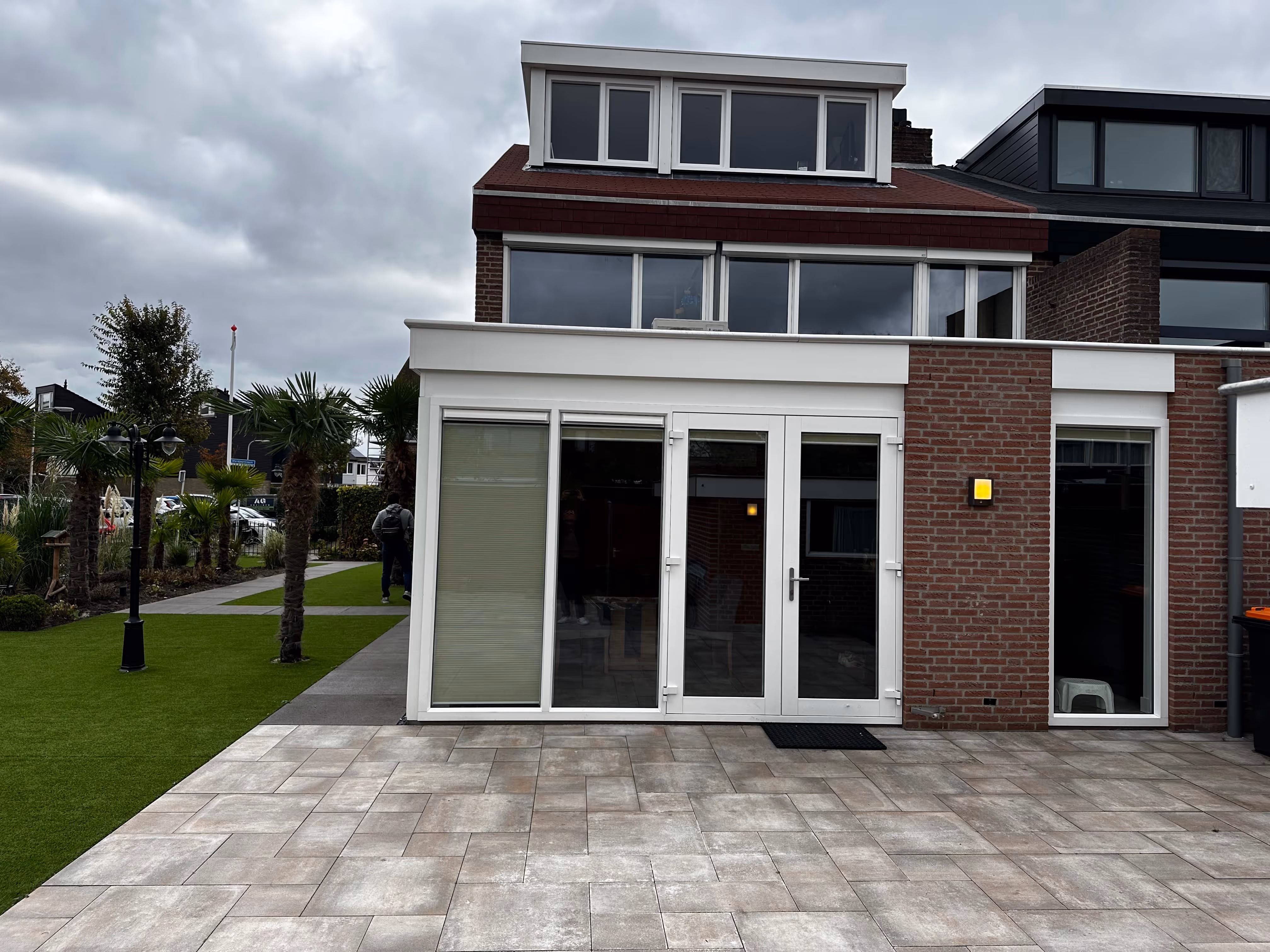 Vooraanzicht van een modern huis met grote ramen, een betegeld terras en palmbomen langs een grasrijke stoep onder een bewolkte lucht.