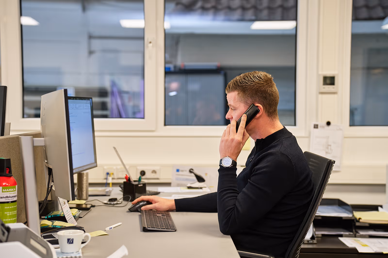 Man die aan een bureau zit, aan de telefoon en met een computer aan het werk in een kantoorruimte.