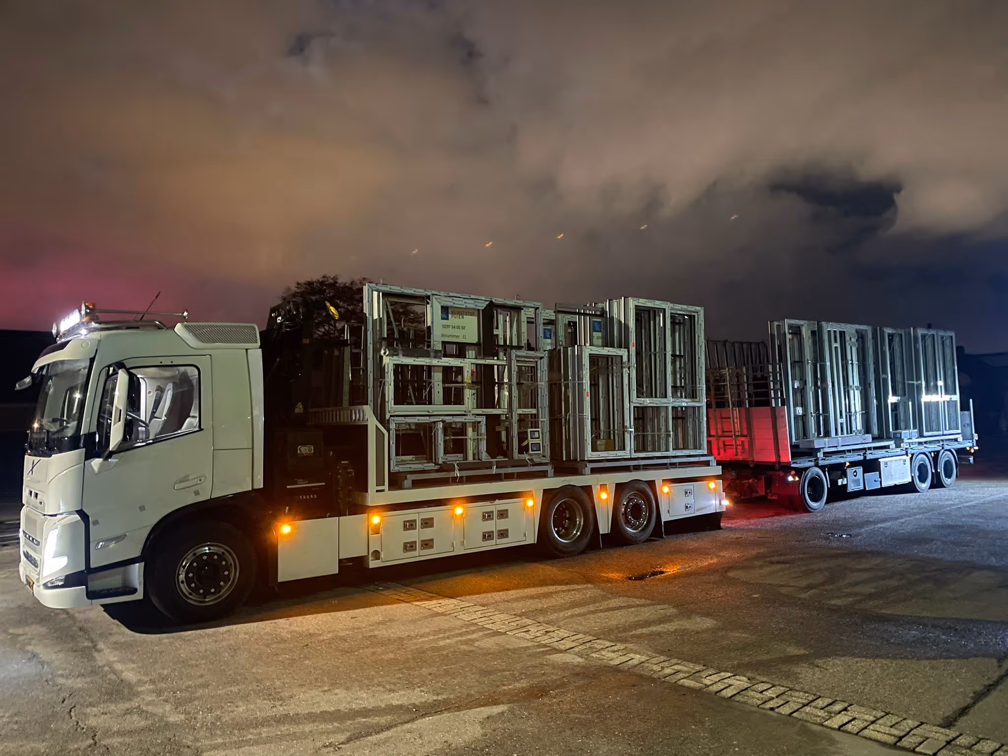 Witte vrachtwagen met aanhanger geladen met metalen raamkozijnen tijdens nachtelijke stop.
