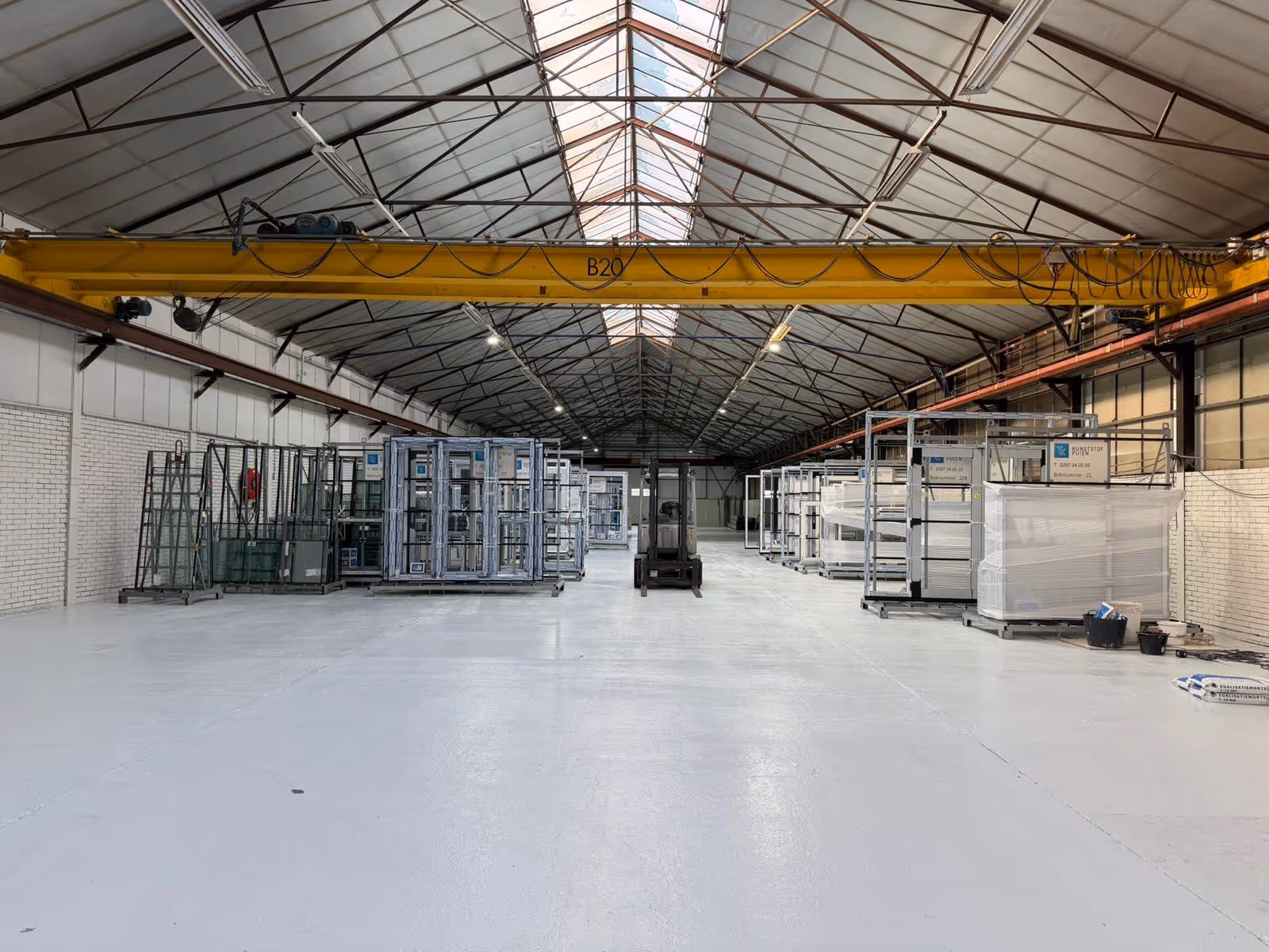 Grote, lege fabriekshal met stapels metalen ramen en deuren en een gele hijskraan aan het plafond.