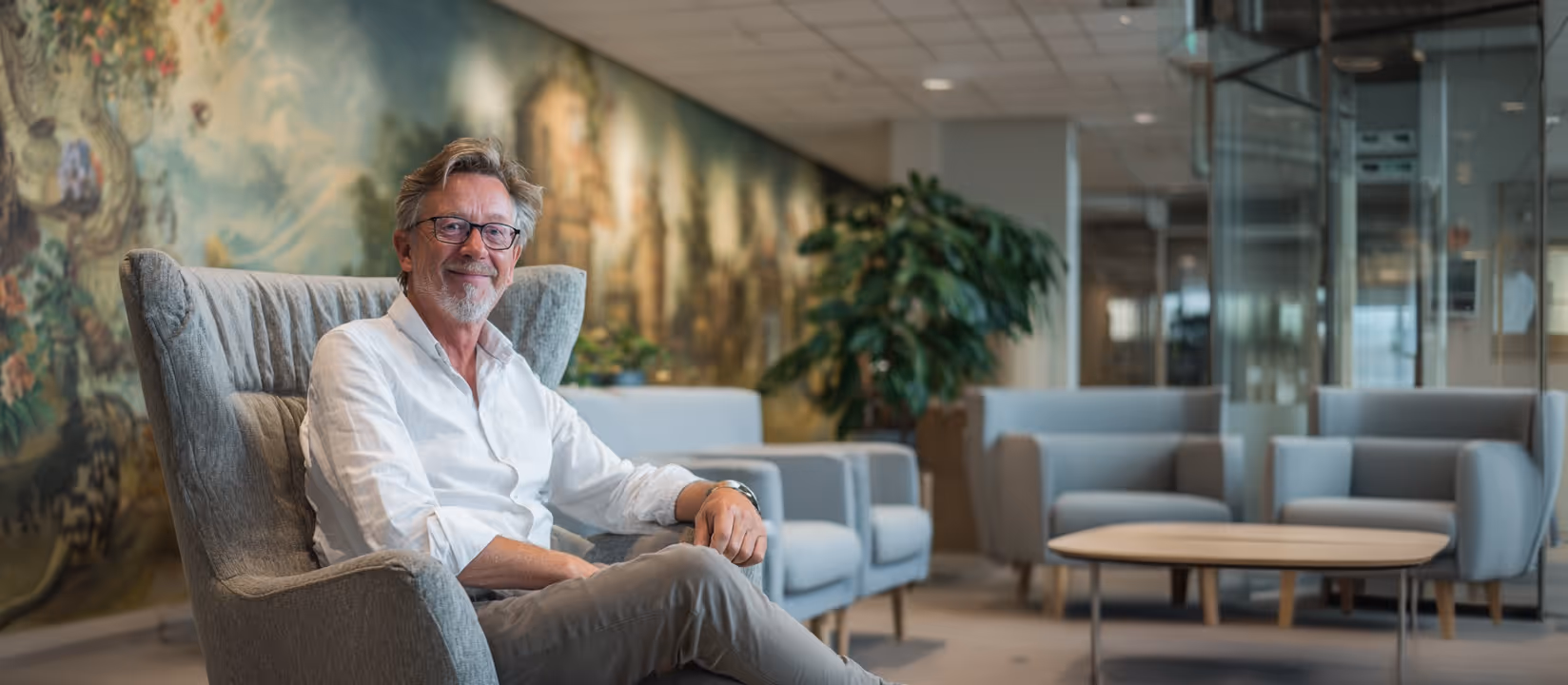 Oudere man met bril zittend in een comfortabele stoel in een moderne wachtruimte met verzachte kleuren en een grote plant.