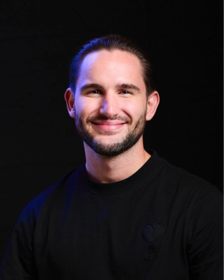 Smiling man with dark hair and beard wearing a black shirt against a black background.