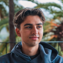 Young man with dark hair wearing a blue hoodie outdoors with palm trees in the background.