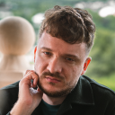 Man with curly hair and beard resting his face on his hand, looking thoughtful indoors.