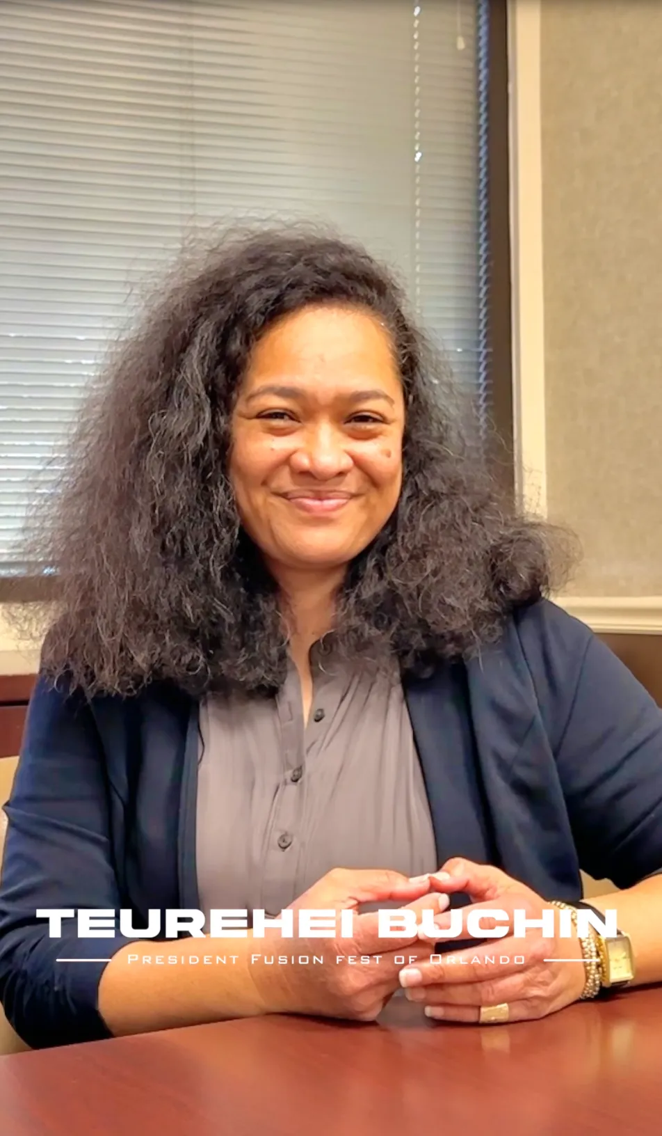 Woman with curly hair smiling, seated at a table in an office setting.