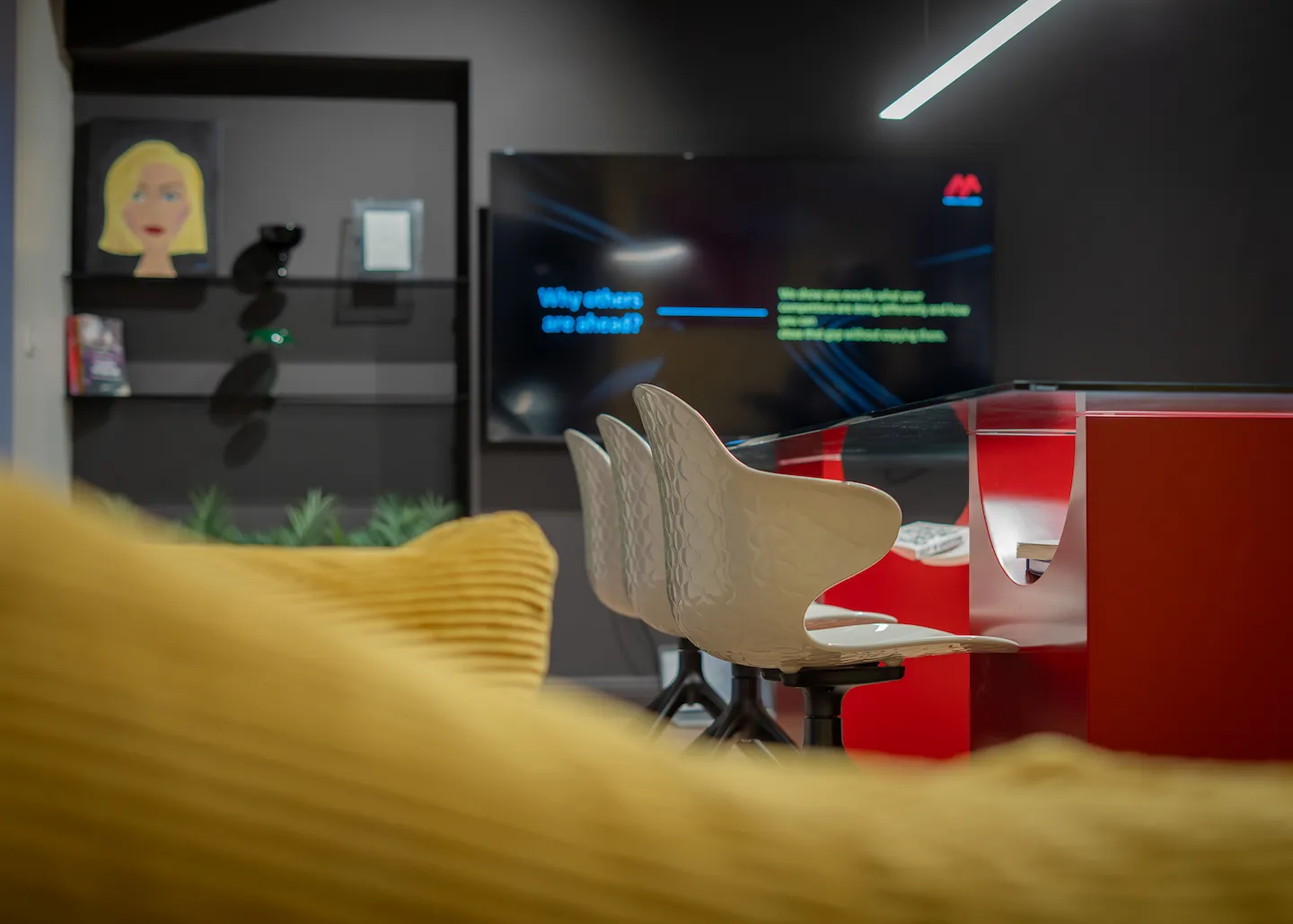 Modern office space with three white swivel chairs at a red table and a TV screen on a dark wall displaying text.