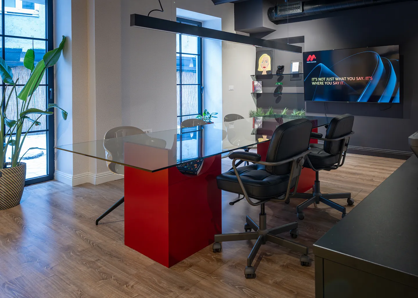 Modern office conference room with a glass table supported by red bases, black leather office chairs, wooden floor, and a large screen displaying a slogan on the wall.