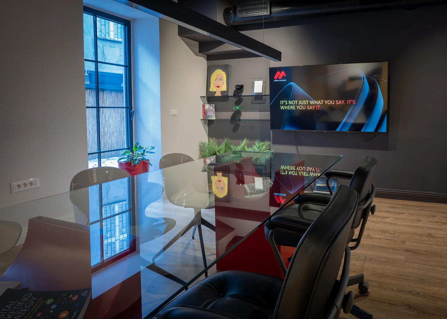 Modern office meeting room with glass table, black chairs, wall-mounted TV displaying the phrase 'It's not just what you say. It's where you say it.'