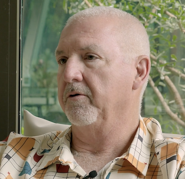 Middle-aged man with short white hair and a trimmed beard wearing a patterned shirt looking slightly to the side, seated near a window with green foliage outside.