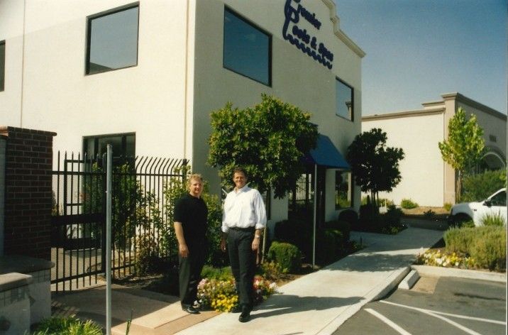 Two men standing on a sidewalk in front of a beige commercial building with trees and flowers around.