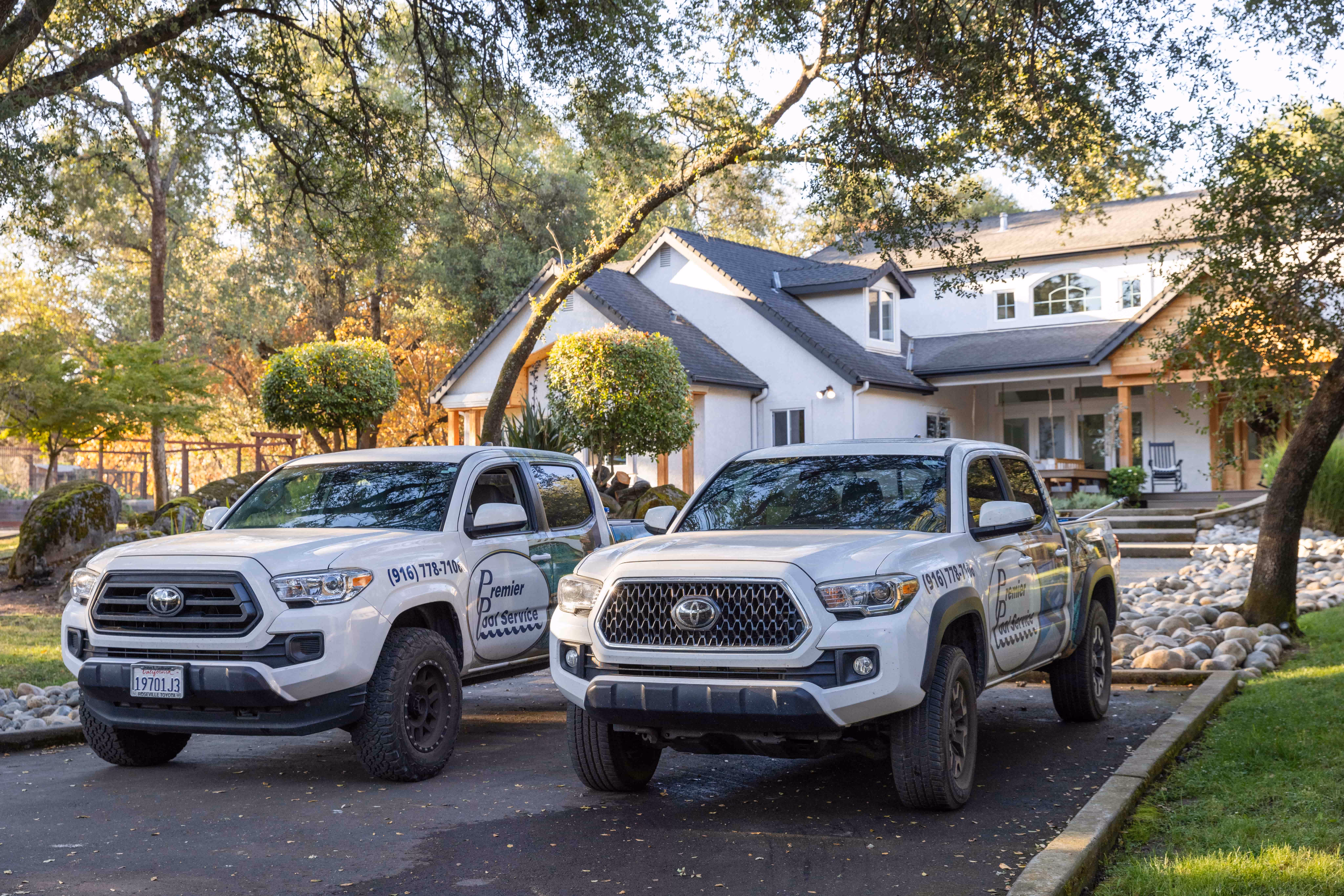 Two white Toyota pickup trucks with Premier Pool Service logos parked on a driveway in front of a large house surrounded by trees and landscaping.
