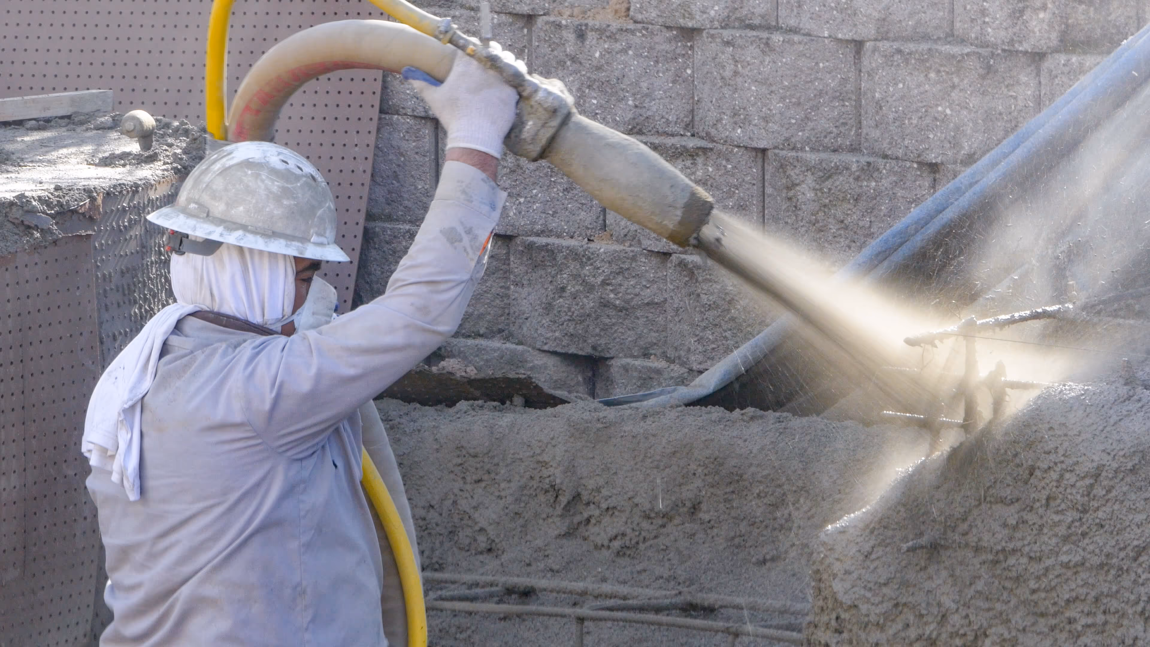 Construction worker wearing a helmet spraying wet concrete onto a wall with a hose.