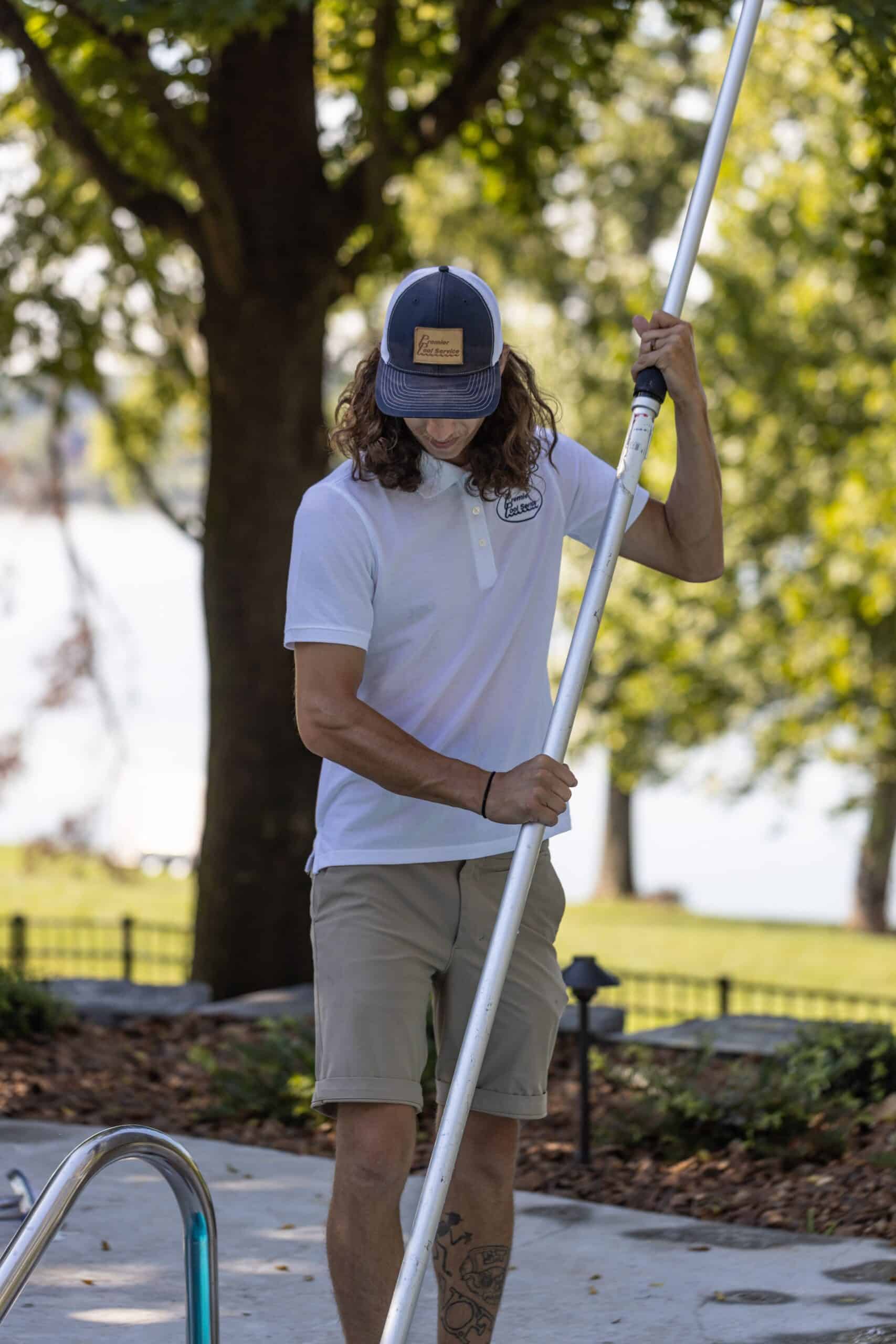 Man wearing a cap and white polo shirt cleaning a swimming pool with a long pole near trees and grass.