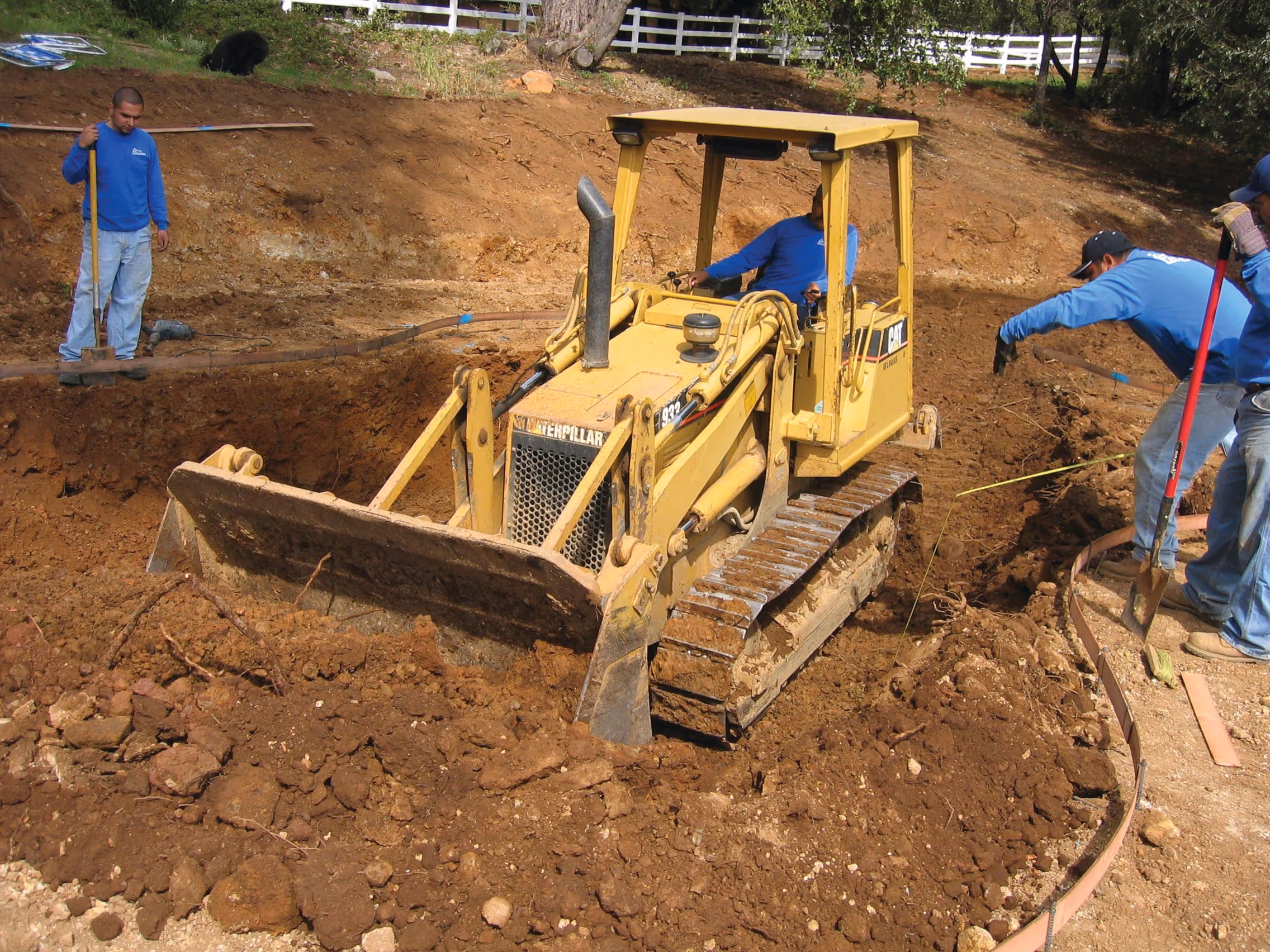 Yellow Caterpillar bulldozer moving earth at a construction site with three workers in blue shirts managing the soil and measuring layout.