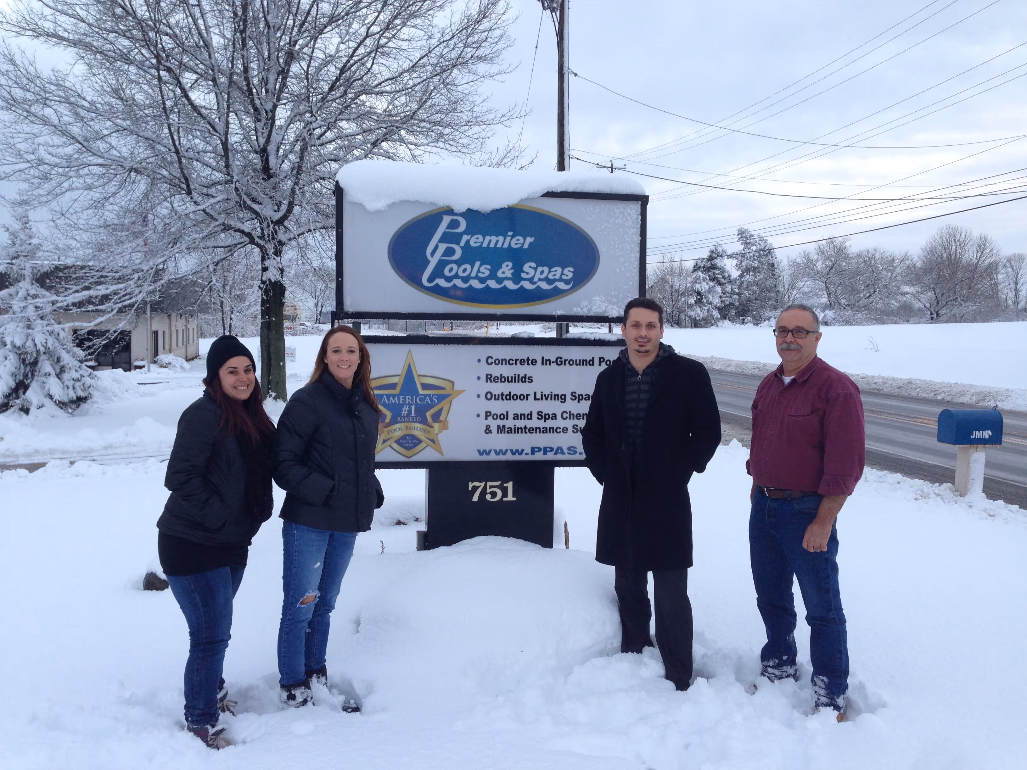 Four people standing in deep snow in front of a Premier Pools & Spas sign during winter.