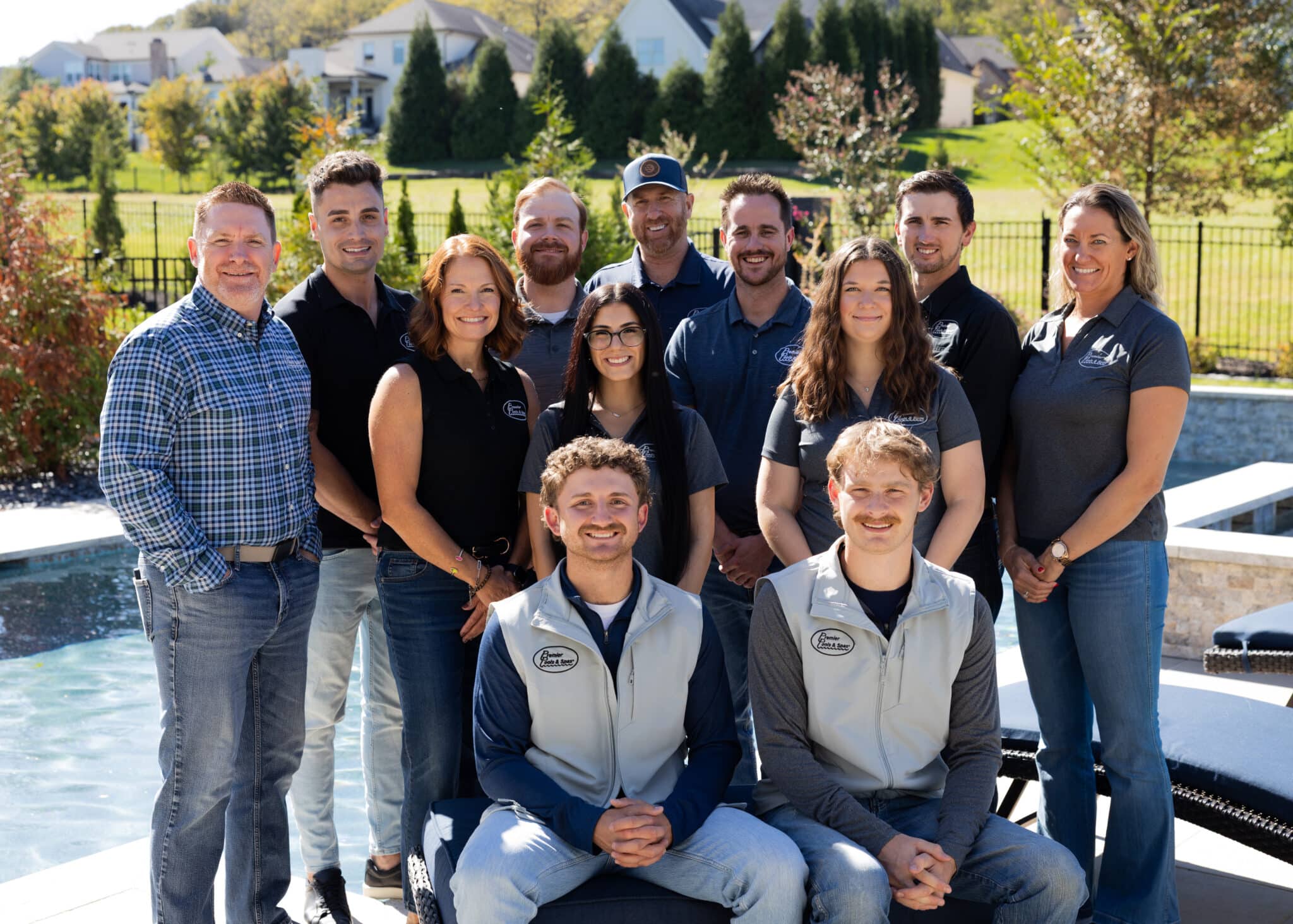 Group of eleven people posing outdoors near a pool with houses and greenery in the background, most wearing branded black or gray shirts and two men seated in front wearing light gray vests.