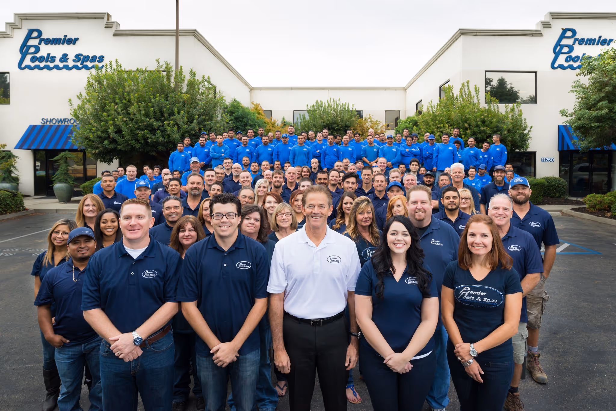 Large group of Premier Pools & Spas employees posing in front of the company building, wearing branded blue and white shirts.