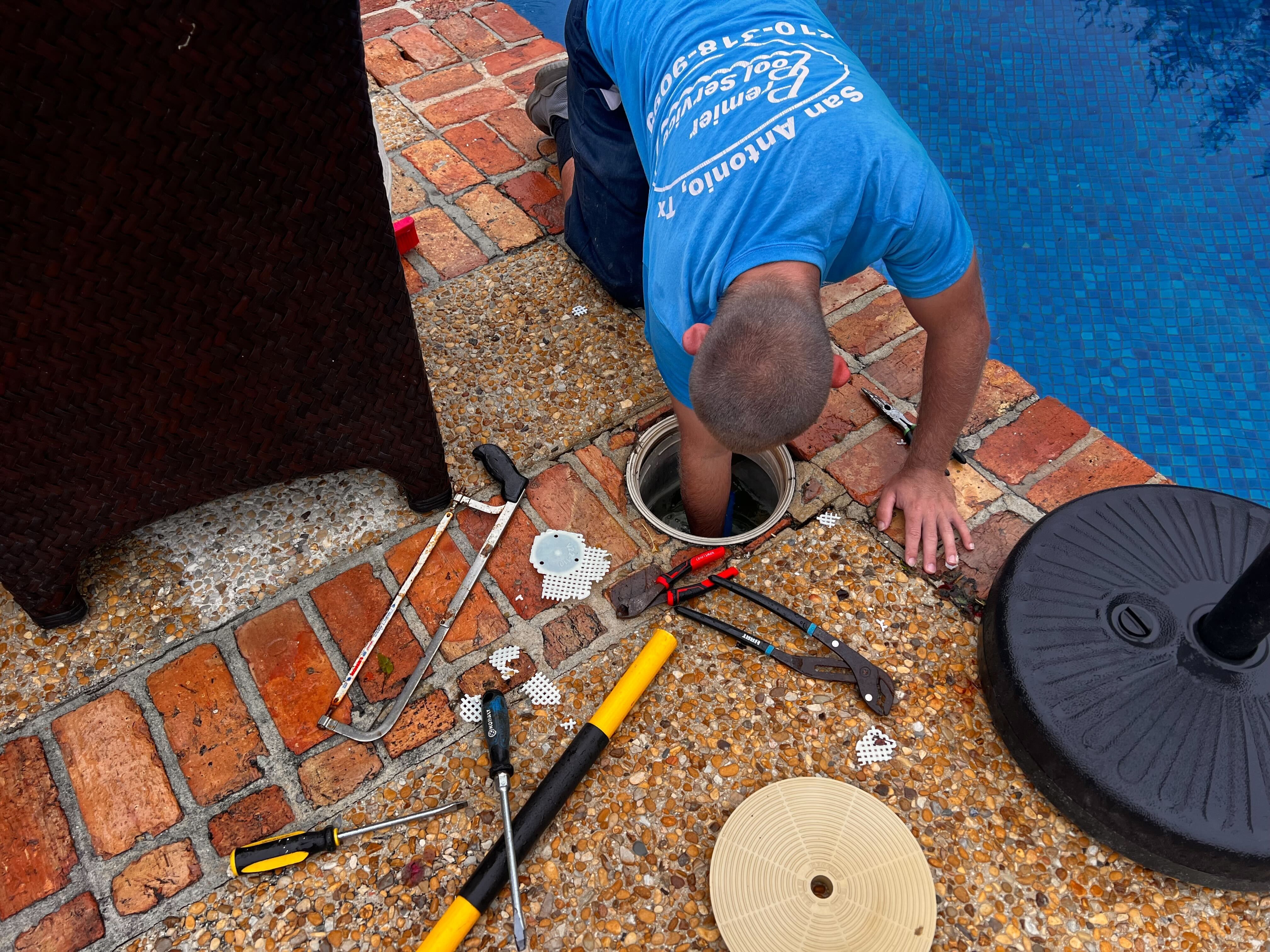 Man in a blue shirt kneeling by a poolside working inside an open pool skimmer with various repair tools scattered on the ground.