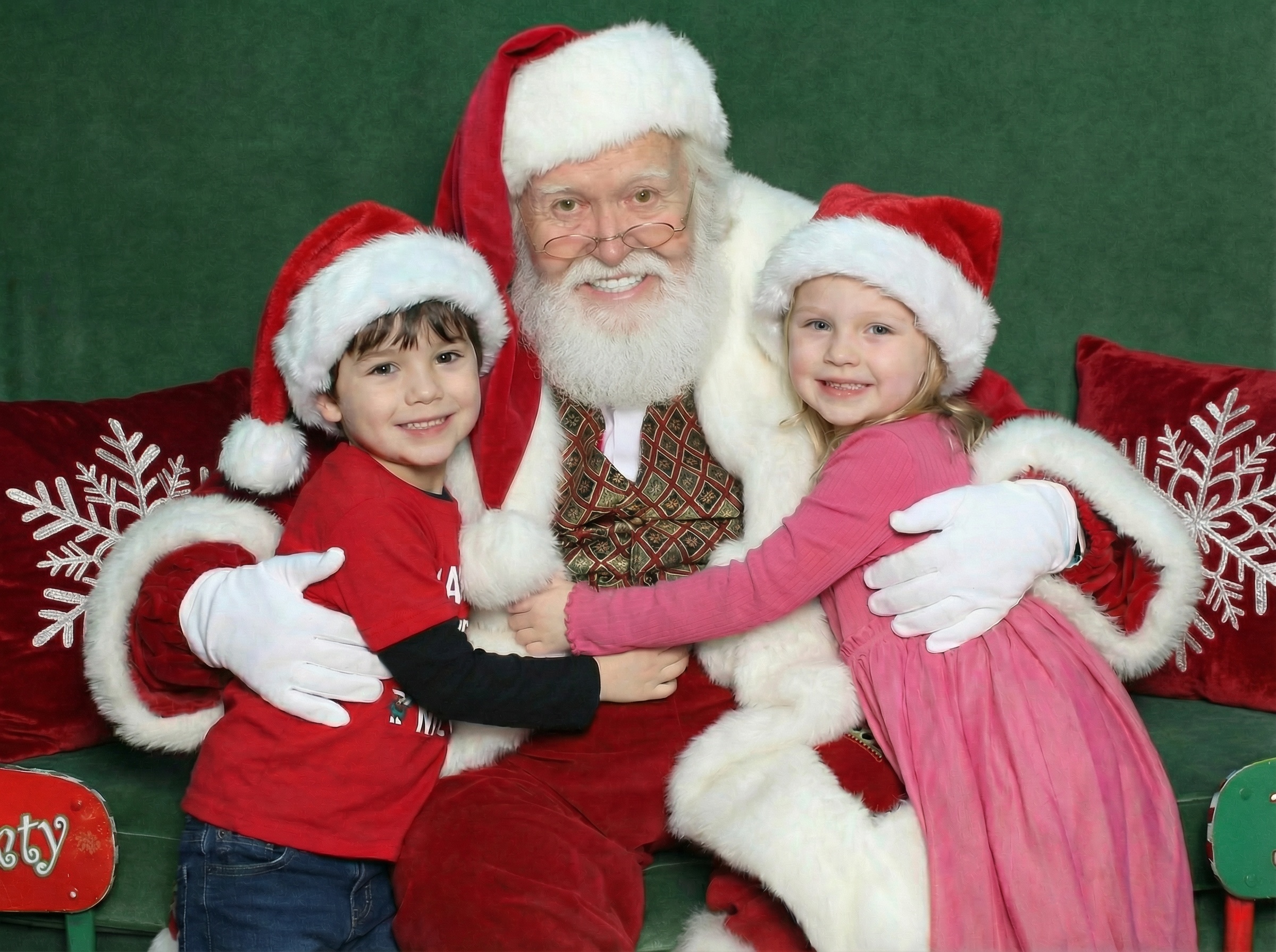 Santa Claus sitting on a green couch with two children, a boy closing his eyes and a girl holding a small sign that says 'Nice'.
