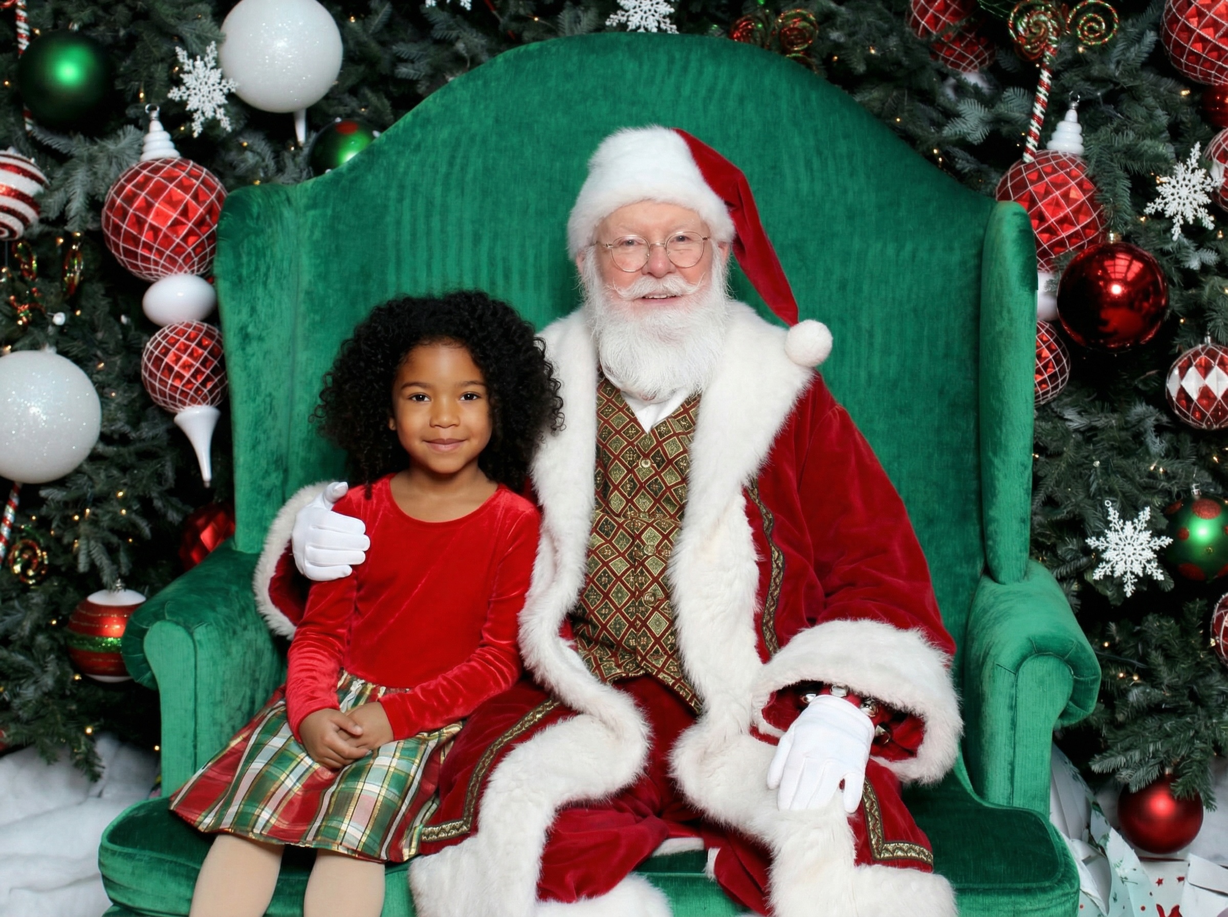 Santa Claus sitting on a green couch with a smiling girl holding a 'Nice' sign and a boy squinting beside him.