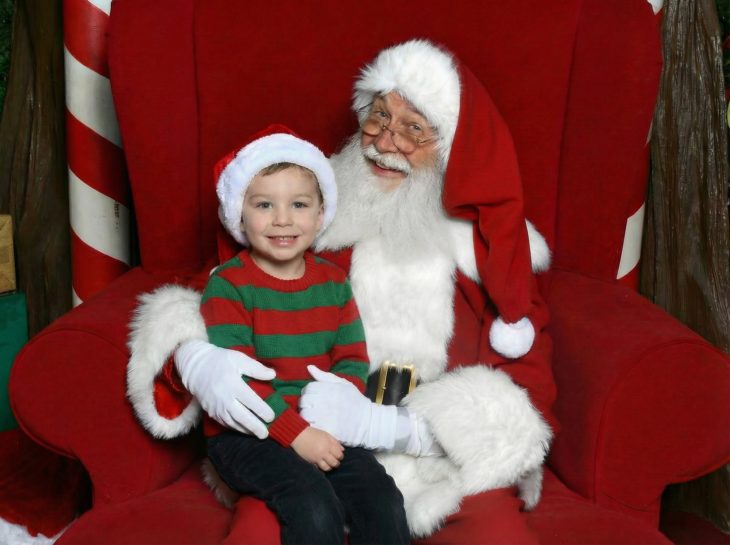 Santa Claus sitting on a green couch with a smiling little girl holding a 'Nice' sign and a boy making a face with his eyes closed.
