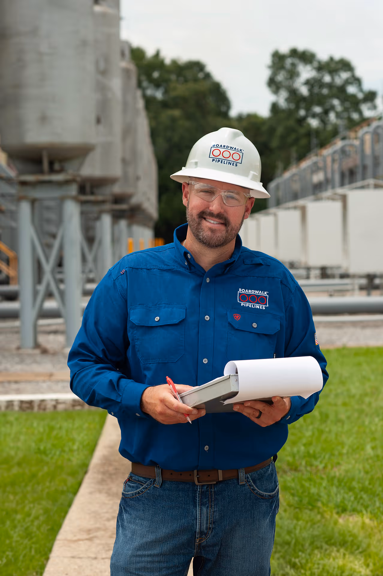 A happy Boardwalk employee wearing a white hardhat, holding a clipboard, and standing in front of pipeline equipment