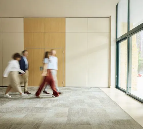 Blurred image of four people walking past a beige wall with wood paneling and large windows.