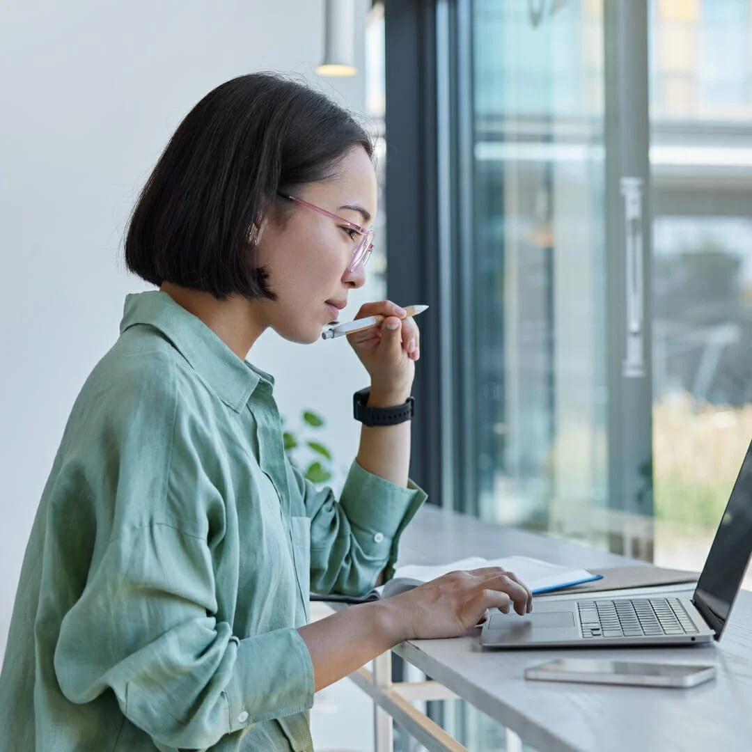 Woman wearing glasses and a green shirt thoughtfully working on a laptop by a window table with a smartphone nearby.