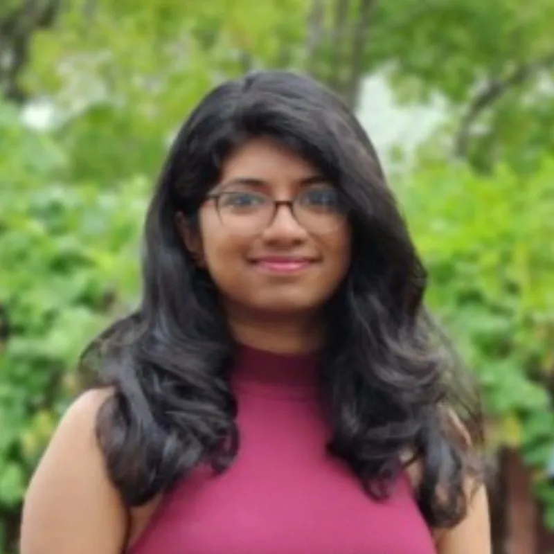 Portrait of a smiling woman with long black hair and glasses wearing a sleeveless maroon top, with a green leafy background.