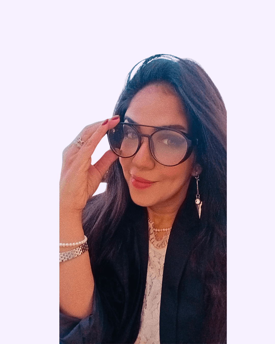 Woman with long dark hair wearing large glasses, a black jacket, pearl necklace, and decorative earrings, smiling and touching her glasses.