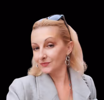 Portrait of a woman with blonde hair wearing a gray coat, silver hoop earrings, and sunglasses on her head, against a black background.