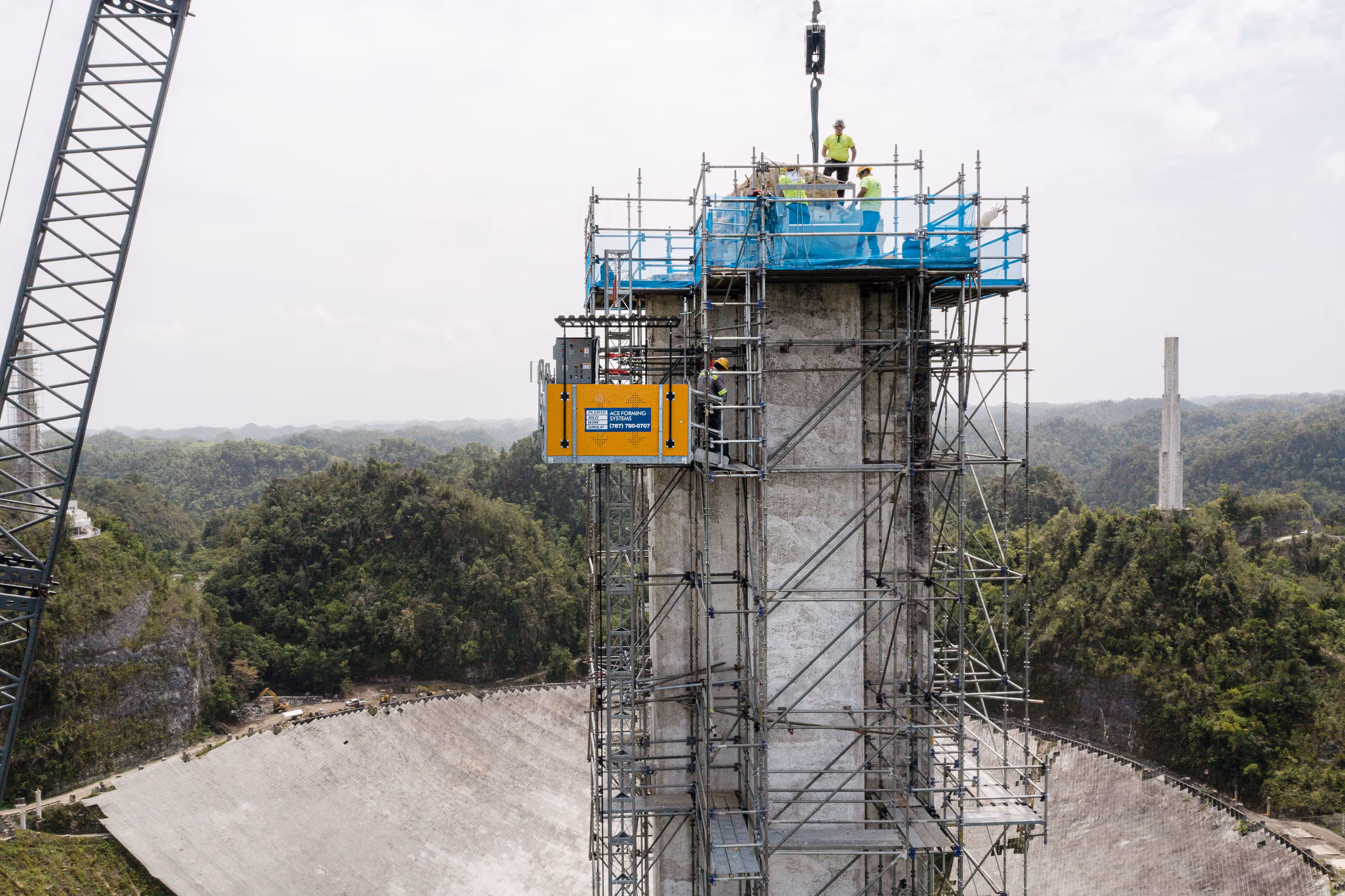 Construction workers atop a scaffolding structure working on a tall concrete pillar surrounded by green forested hills.