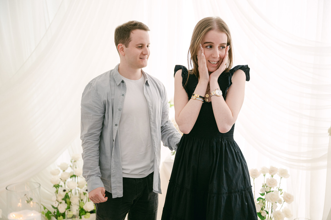 Smiling man looks at a woman in a black dress who is holding her face in surprise, standing together indoors in front of a light curtain.